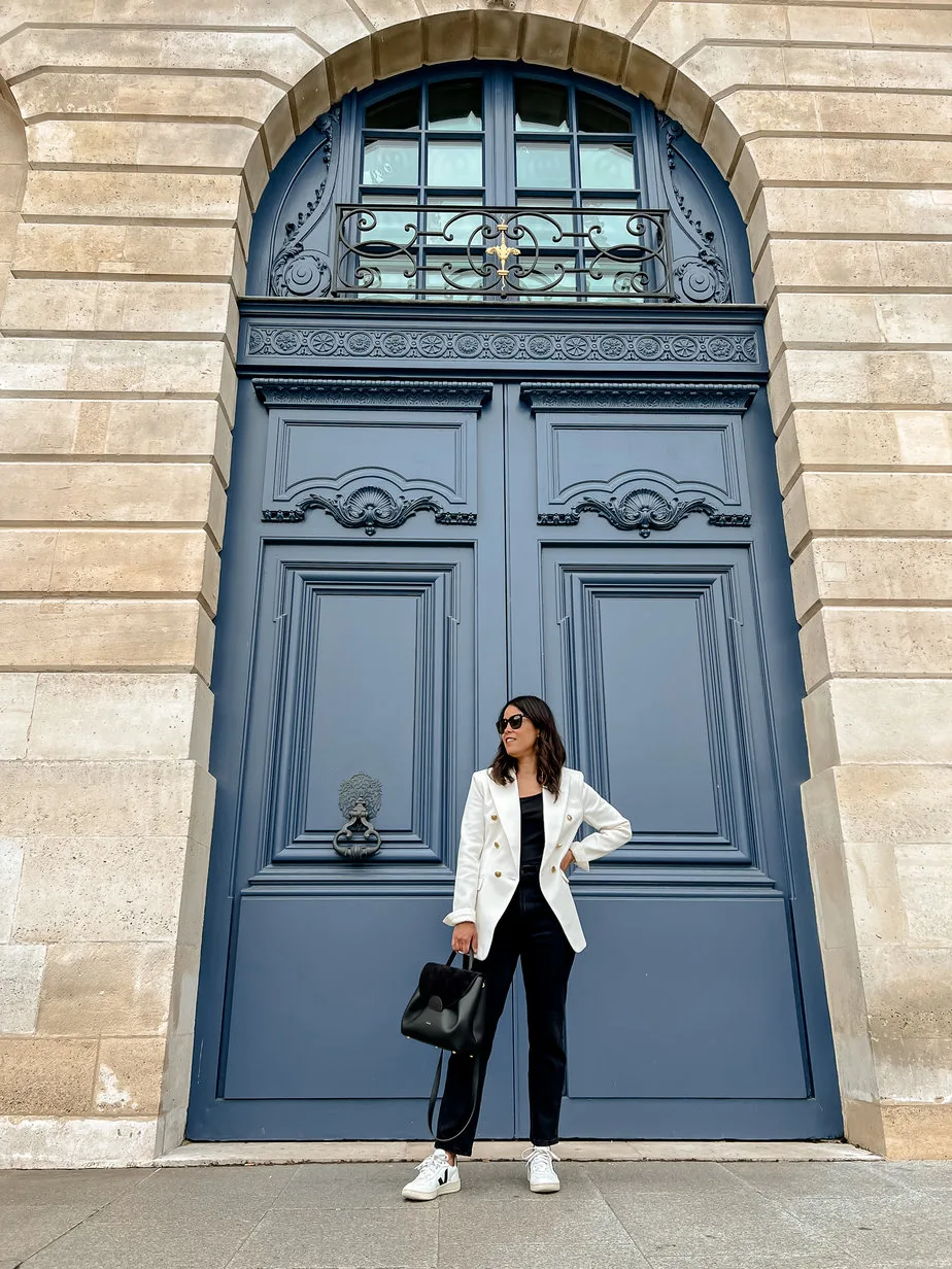Wearing a white blazer, black jeans, veja sneakers and carrying black purse in front of blue door in Paris