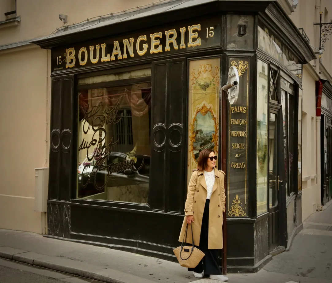 WOMAN WITH TRENCH COAT, MARKET BAG STANDING AGAINST A BOULANGERIE BUILDING IN PARIS.