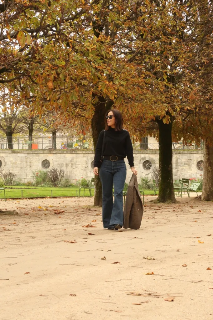 Woman walking wearing jeans and carrying a blazer in a park with trees.