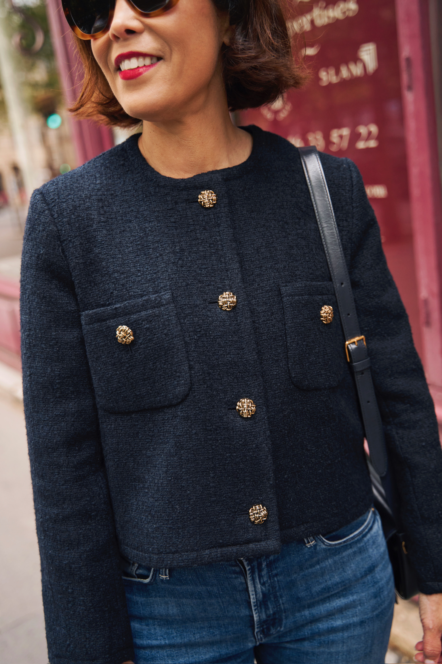 Woman in navy tweed jacket with sunglasses and standing in front of red door in Paris.