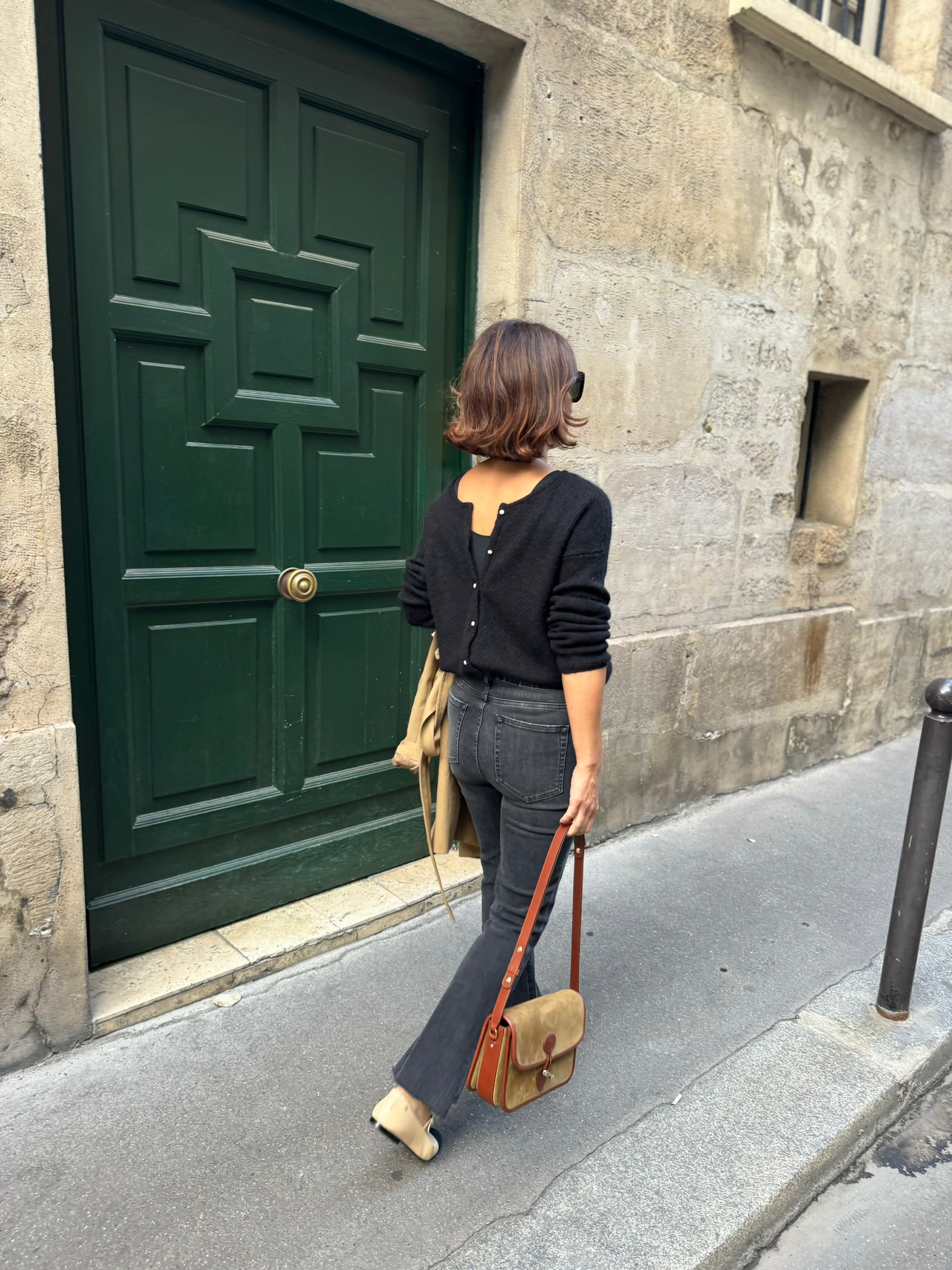Woman wearing black button down sweater with denim and ballet flats walking in Paris in front of a green door.