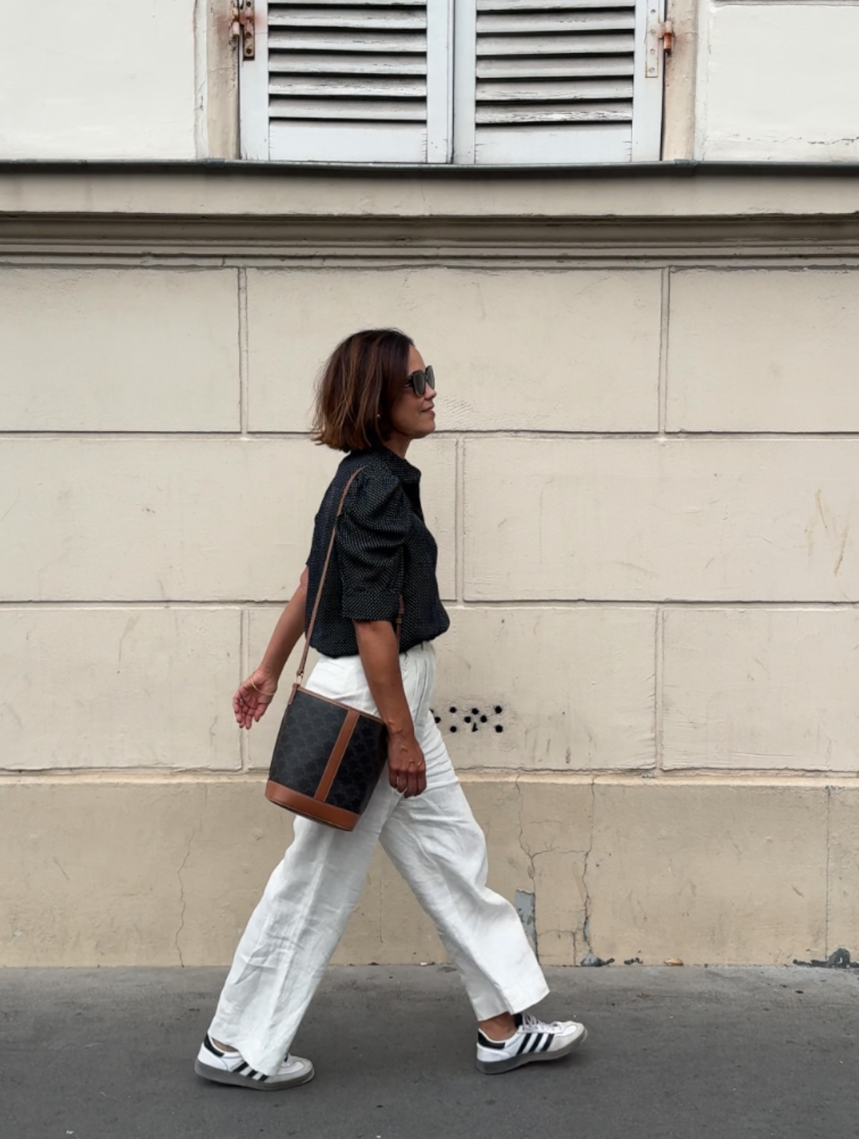 Woman walking against a wall wearing white linen pants, adidas, celine bag, and short sleeve top. 