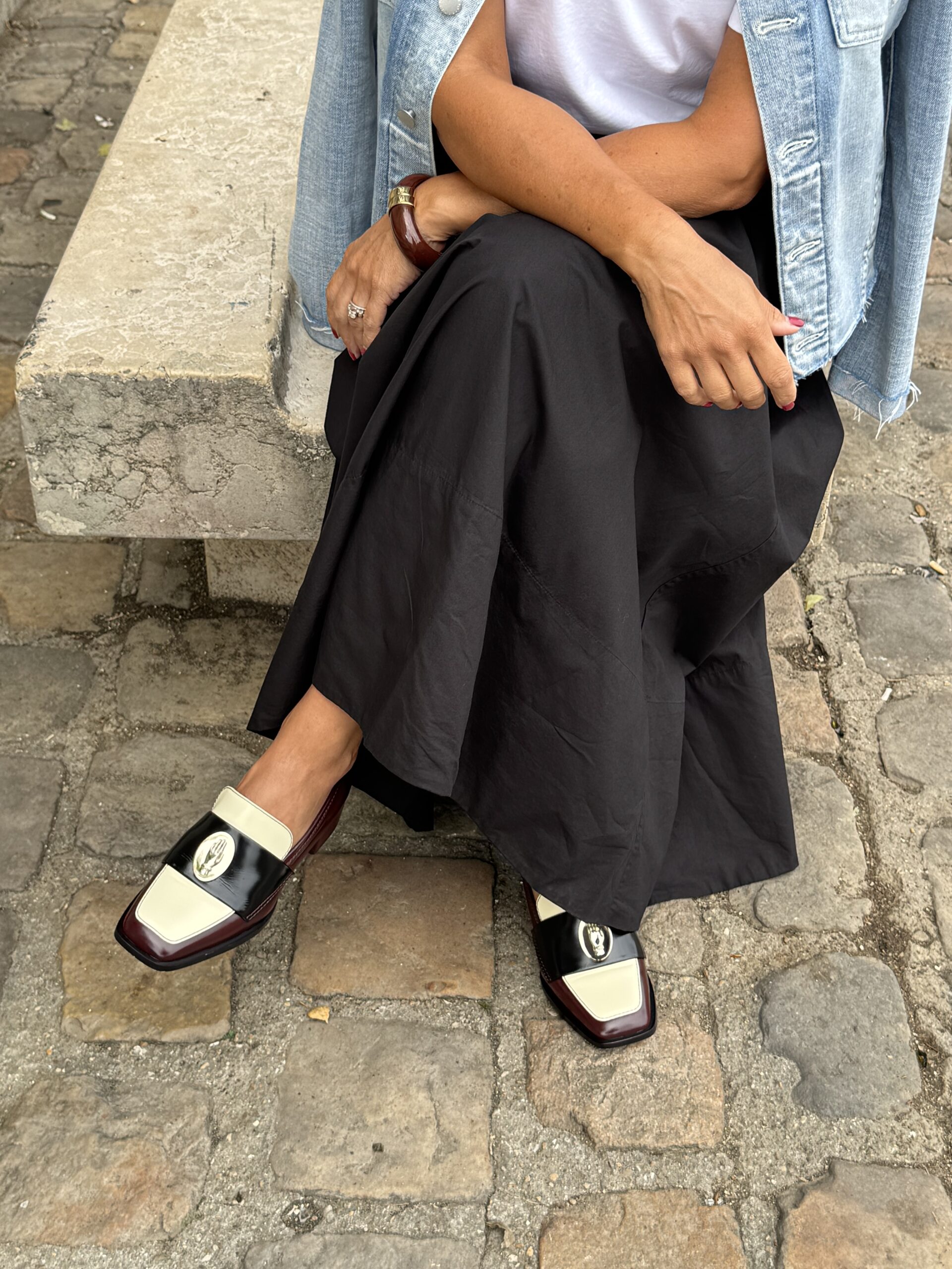 Lady sitting wearing black skirt with loafers on a bench with denim jacket on shoulders.