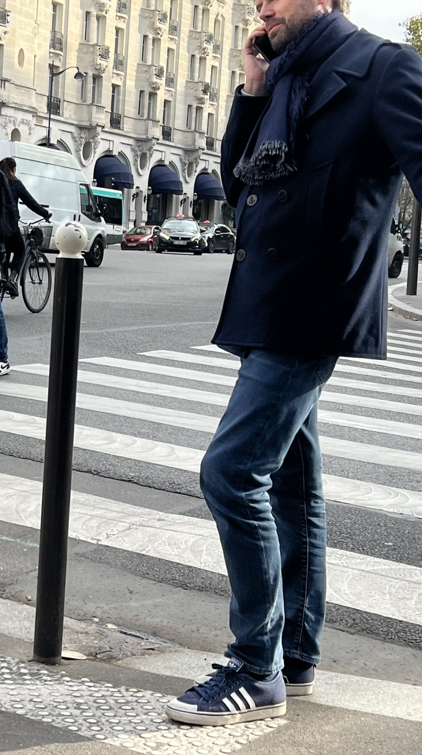 Man wearing navy coat and denim with sneakers standing against pole in Paris.
