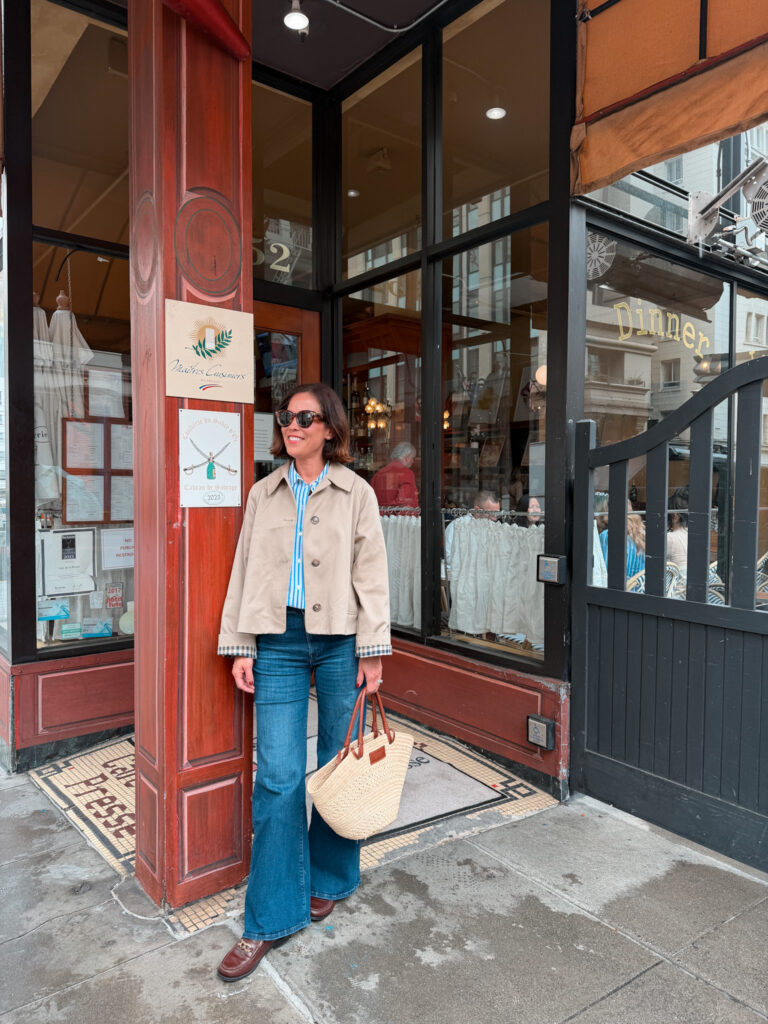 Parisian spring outfit featuring a cropped trench coat styled with denim, a striped blouse, and a straw tote outside a classic café for an effortless French look