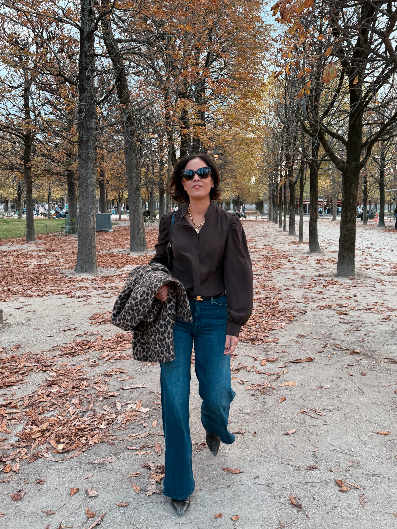 Woman wearing chocolate silk shirt, denim, boots, walking in garden in paris during the fall and autumn seasons.
