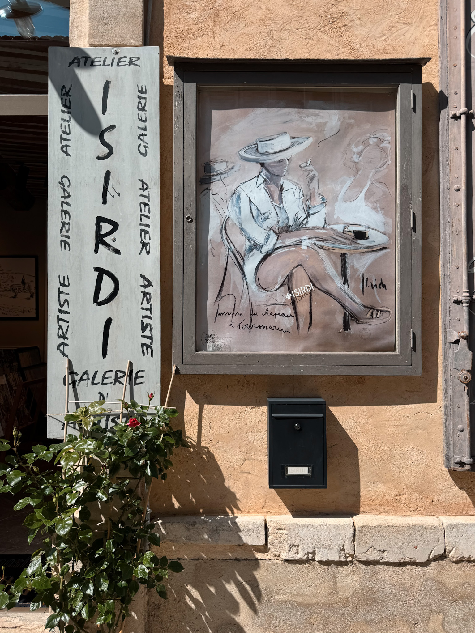 Isirdi Art Gallery storefront with framed artwork on a stone wall in Lourmarin village, Provence