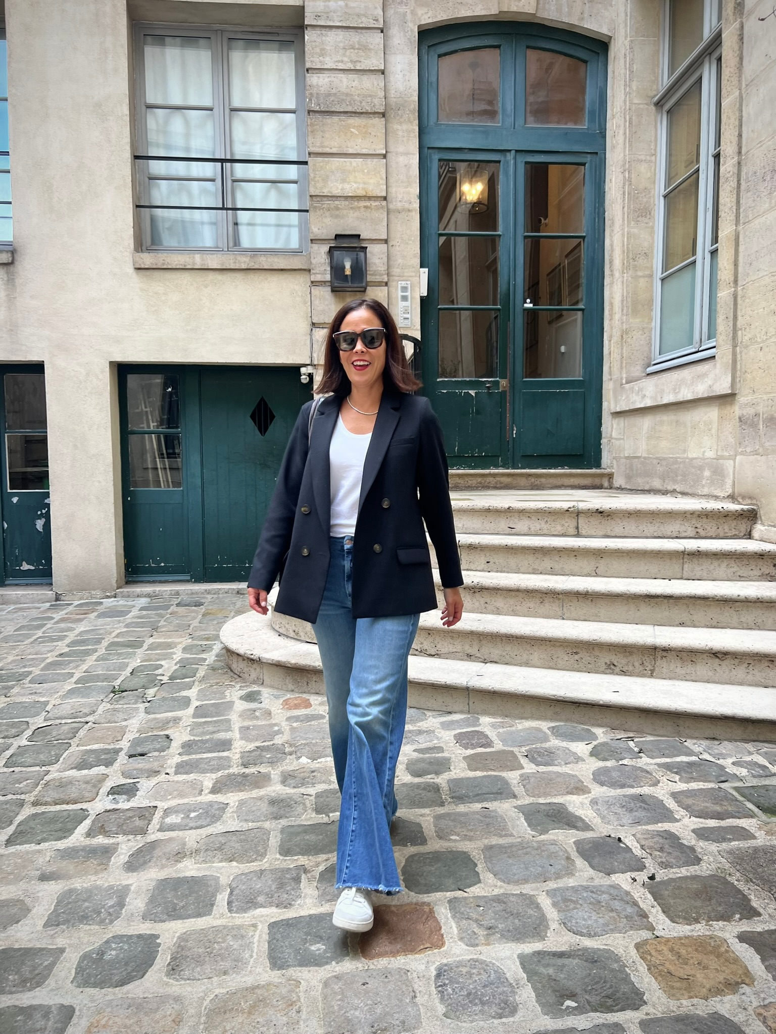 Parisian woman wearing a classic blazer, jeans, and white sneakers in a Paris courtyard