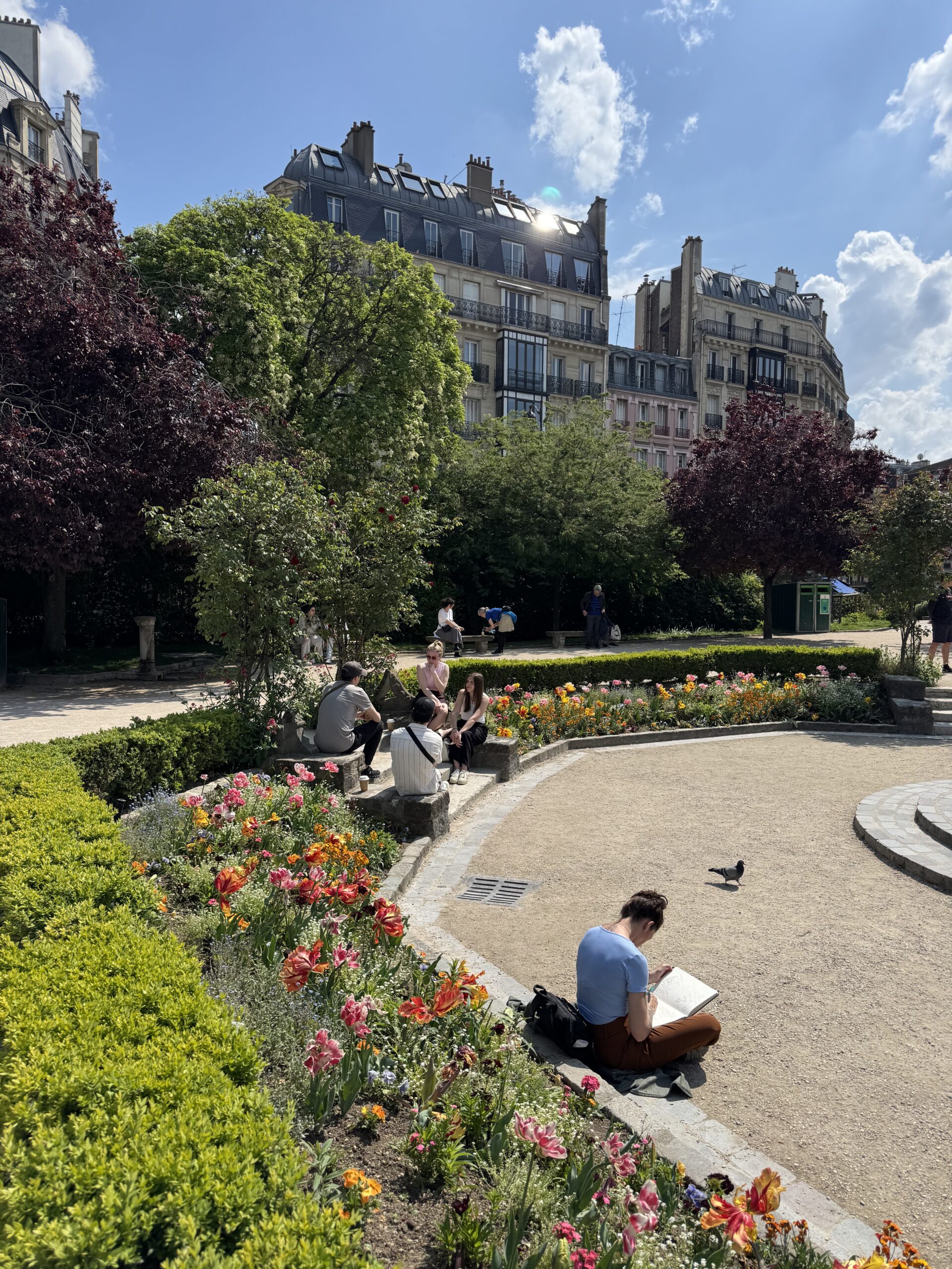 Locals relaxing in a Paris park on a sunny day with buildings in the background.