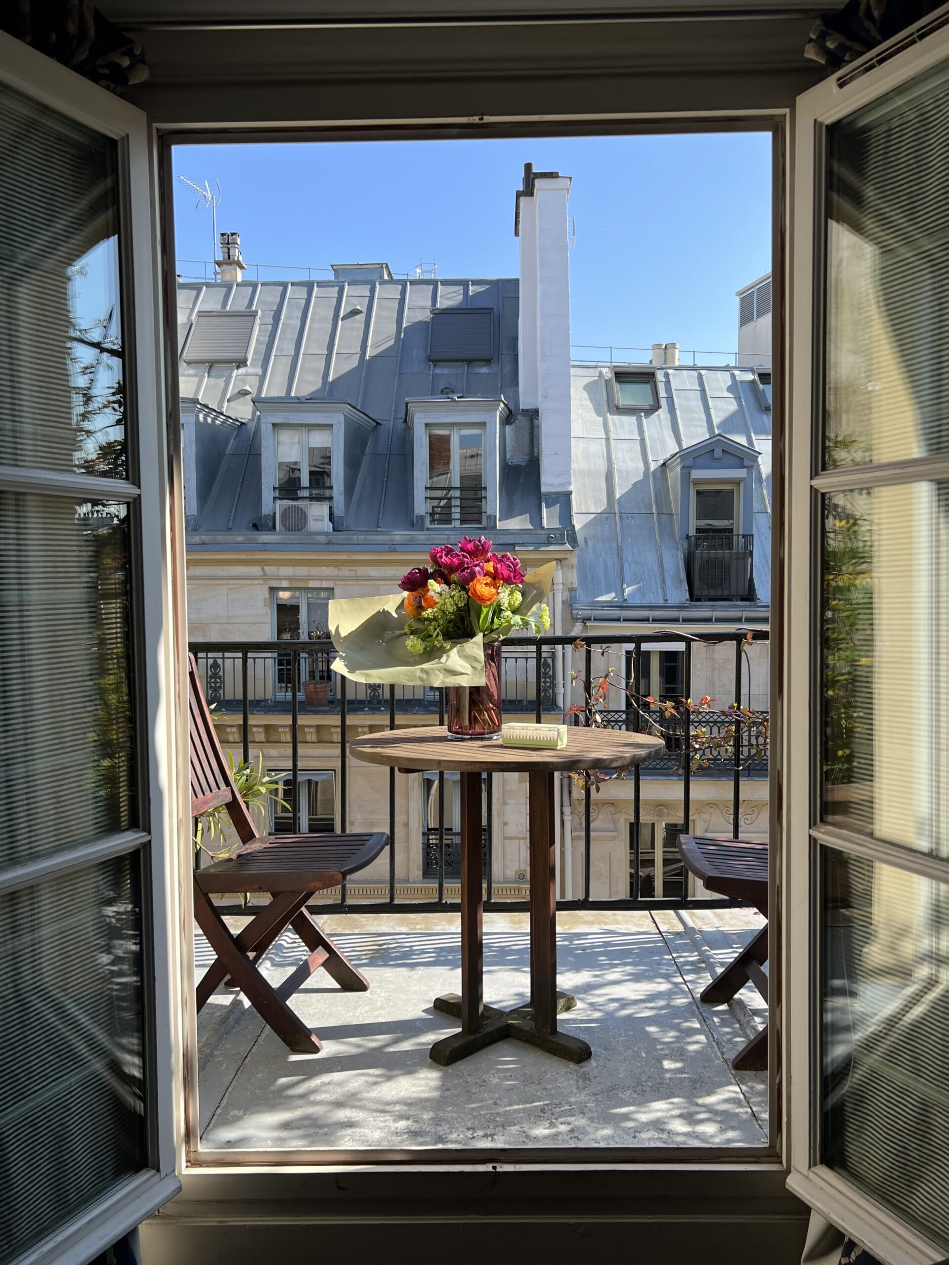 Sunny spring balcony in Paris with Haussman buildings in the background and flowers in a vase on a table.