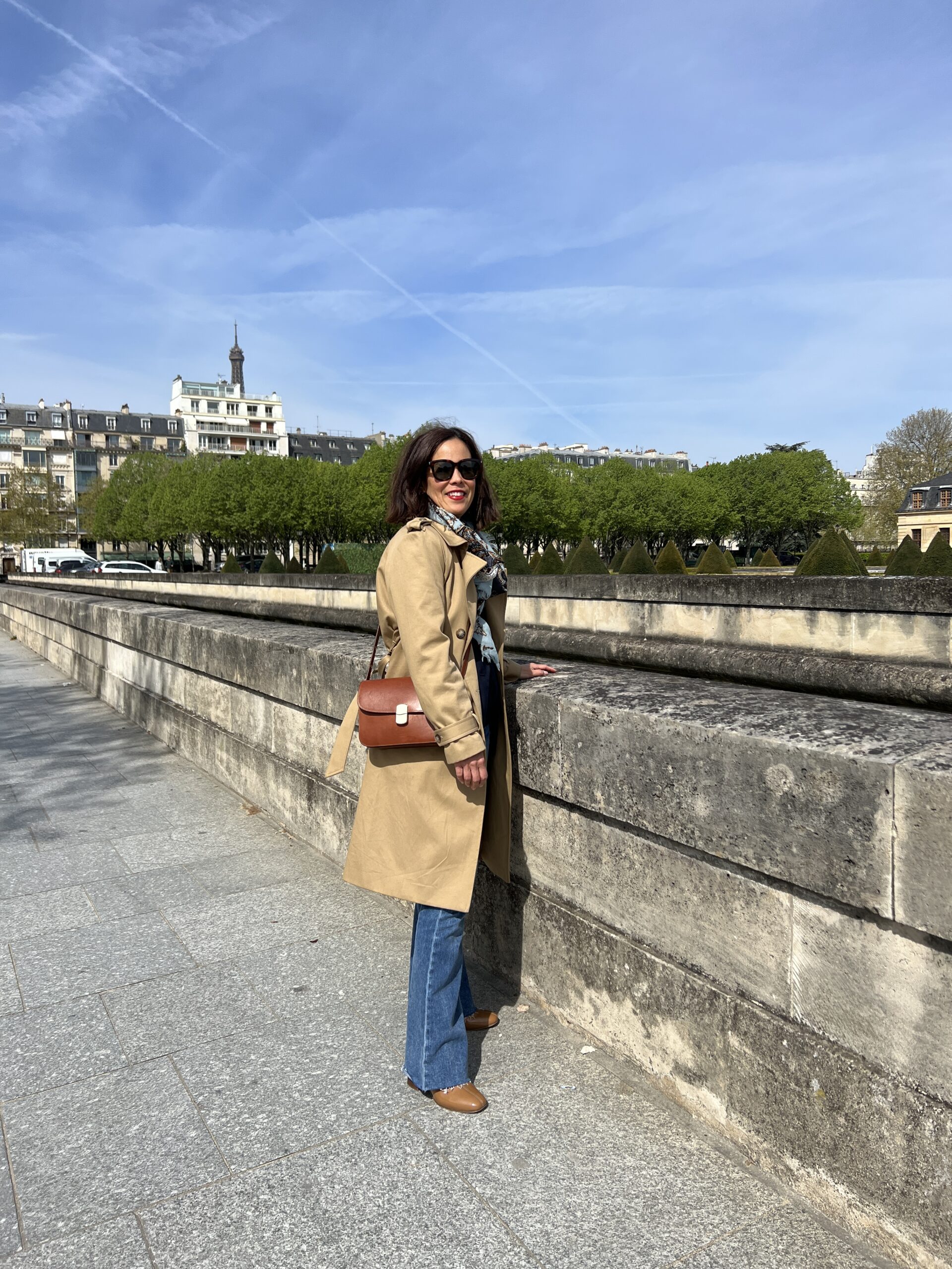Woman wearing a trench coat, crossbody bag, denim, and boots standing in front of a wall.