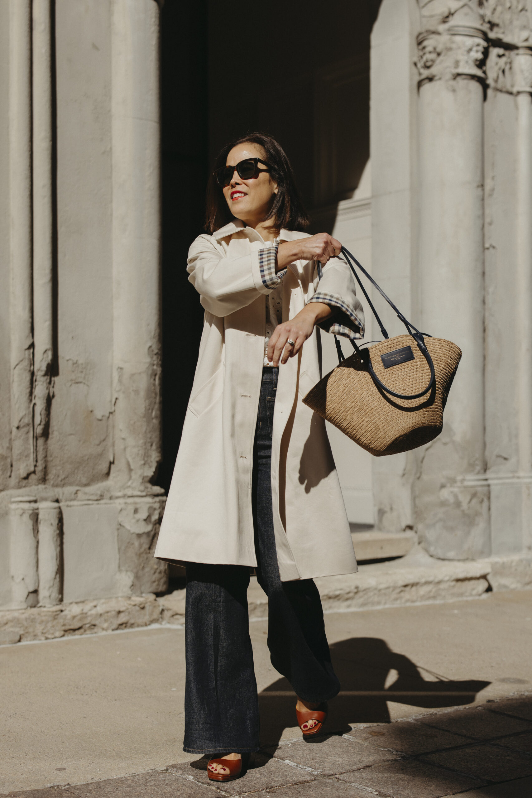 Woman wearing trench coat with eyelet top, denim, basket bag and sezane spring sandals.