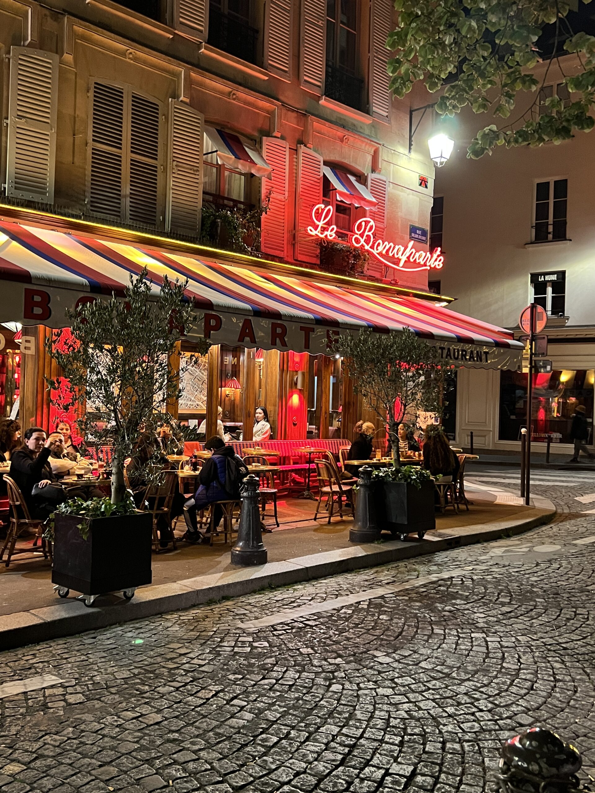 Le Bonaparte Café at night in Saint-Germain-des-Prés with glowing red signage, striped awning, and lively Parisian terrace dining
