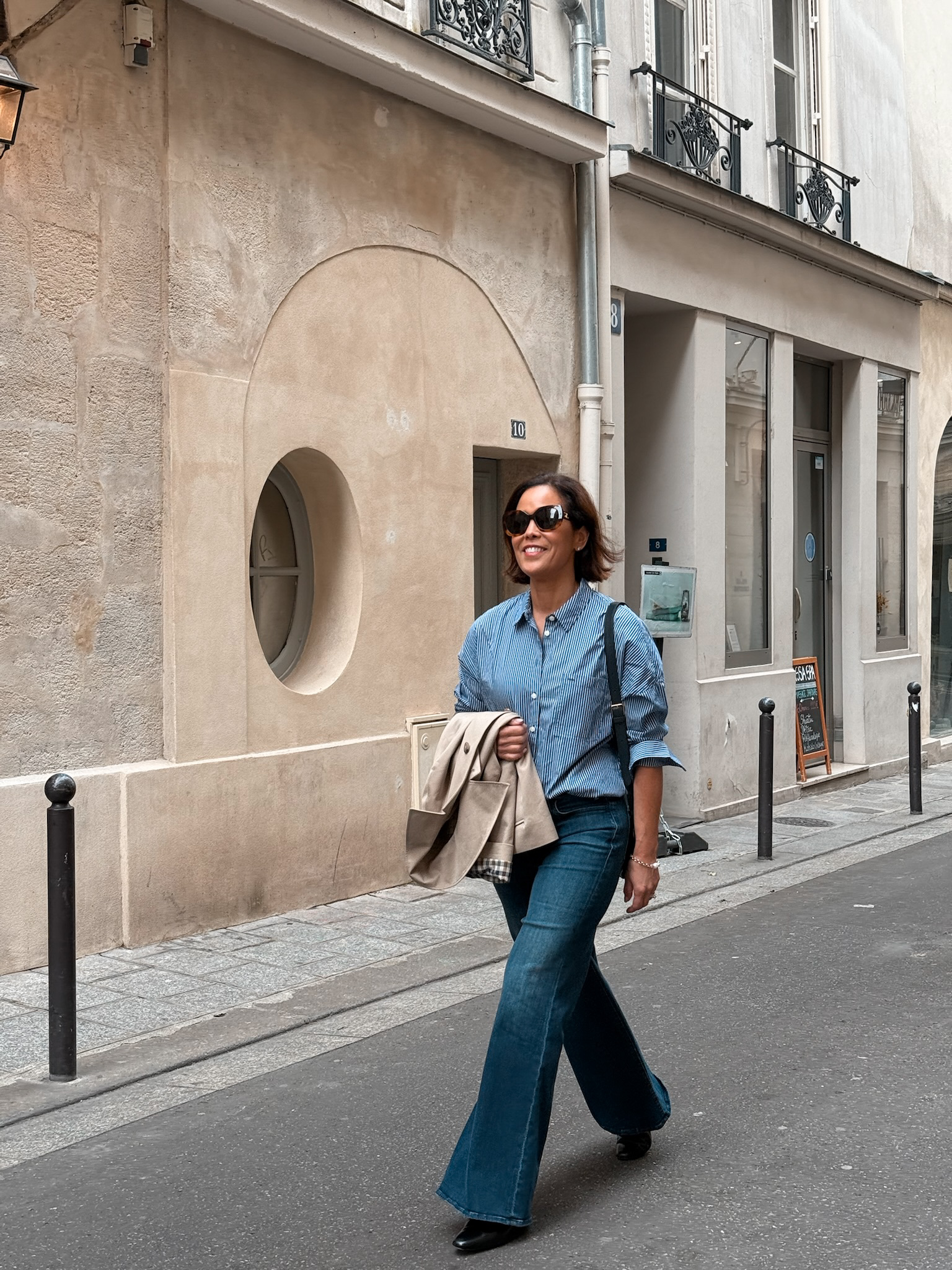 What to wear in Paris in April with trench coat, striped shirt, dark flared jeans, and sunglasses on a Paris street