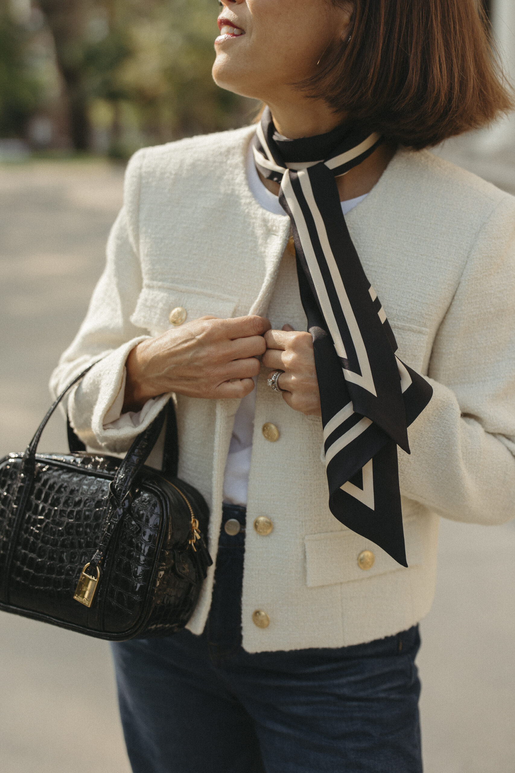 Close-up detail of a cream tweed jacket with gold buttons, a black and cream silk neck scarf, and a black croc-embossed bag — French girl style accessories