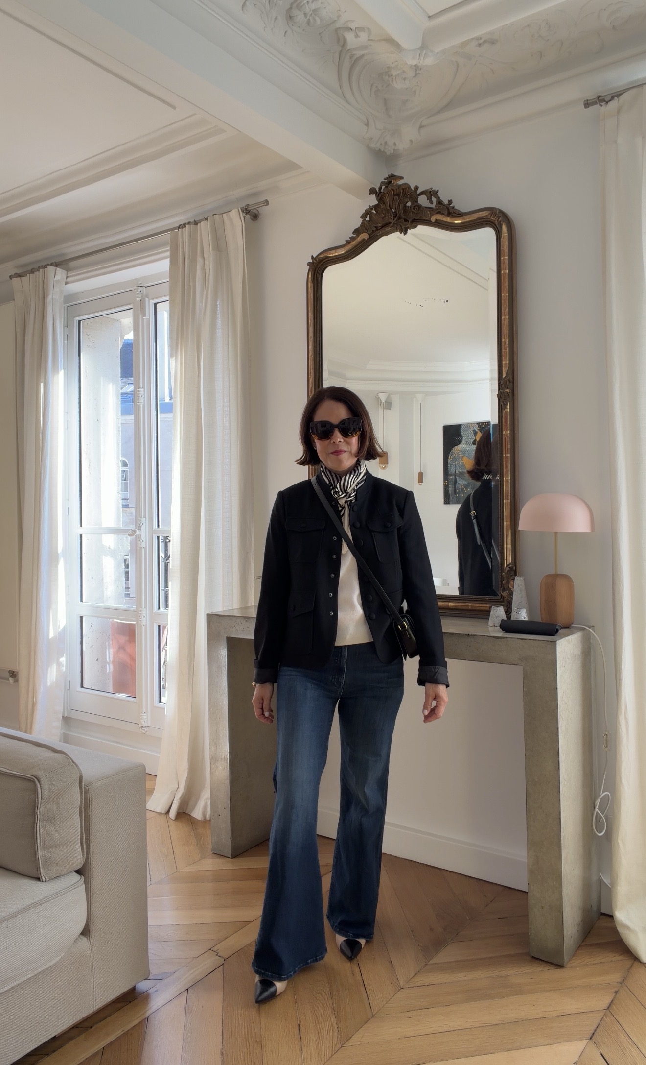 Woman in black utility jacket, cream blouse, flare jeans, and black pointed mules standing in front of gilded mirror in Paris Airbnb
