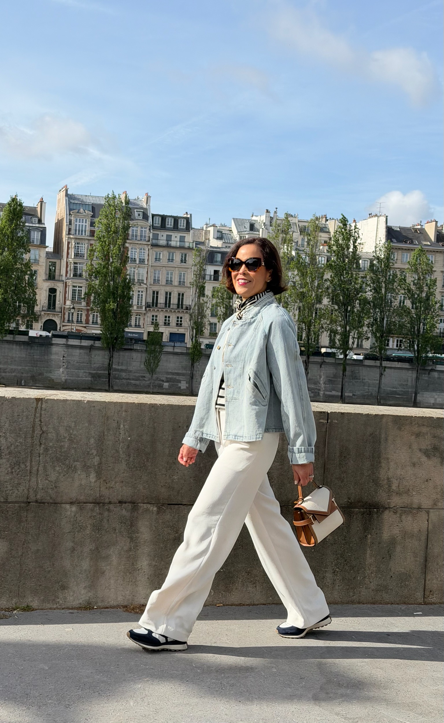 Woman in light denim jacket, ivory wide-leg trousers, and two-tone sneakers walking along the Seine River in Paris