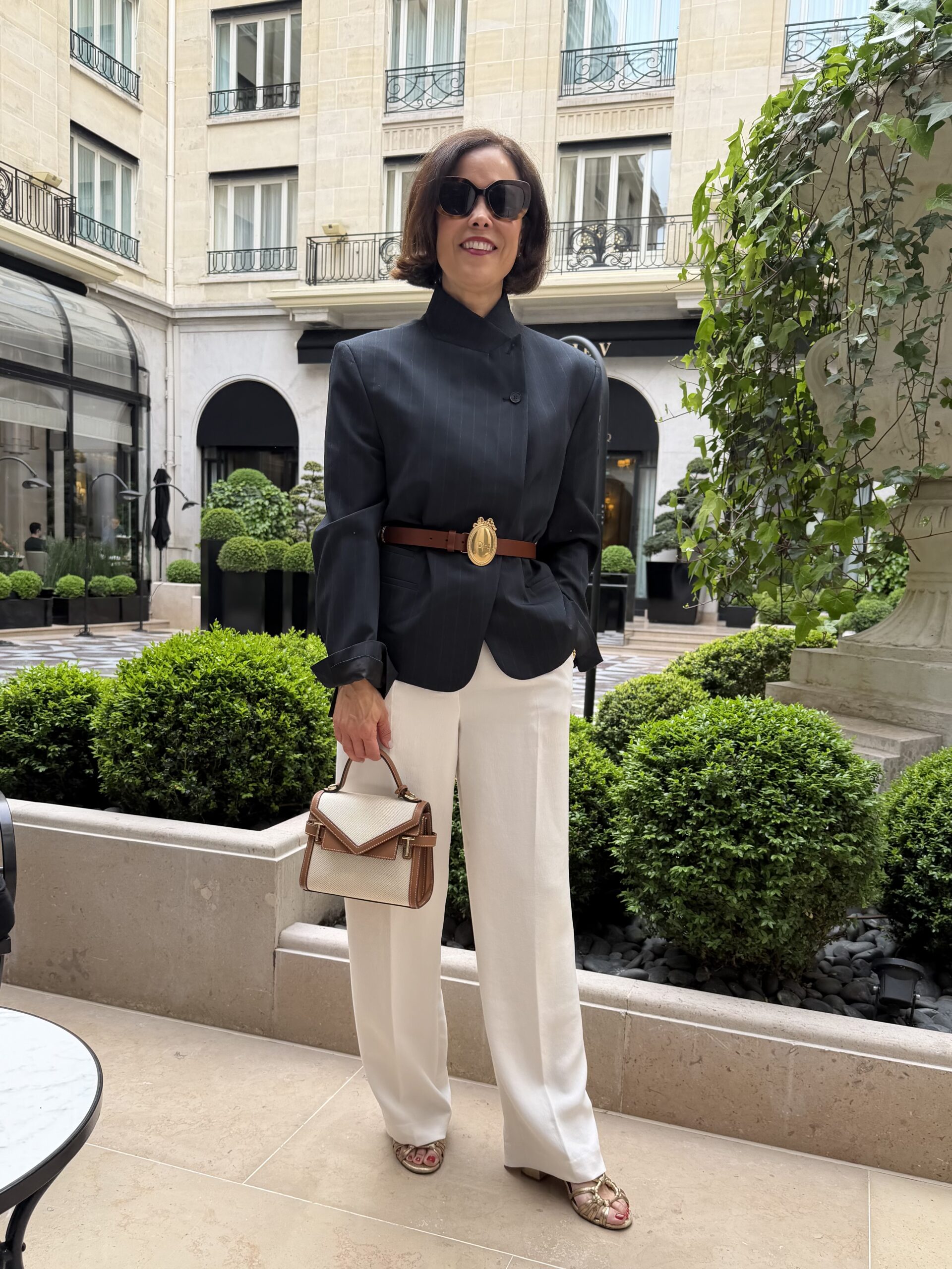 Chic Paris outfit with black blazer, white wide-leg trousers, brown belt, and structured handbag at luxury Paris hotel courtyard.
