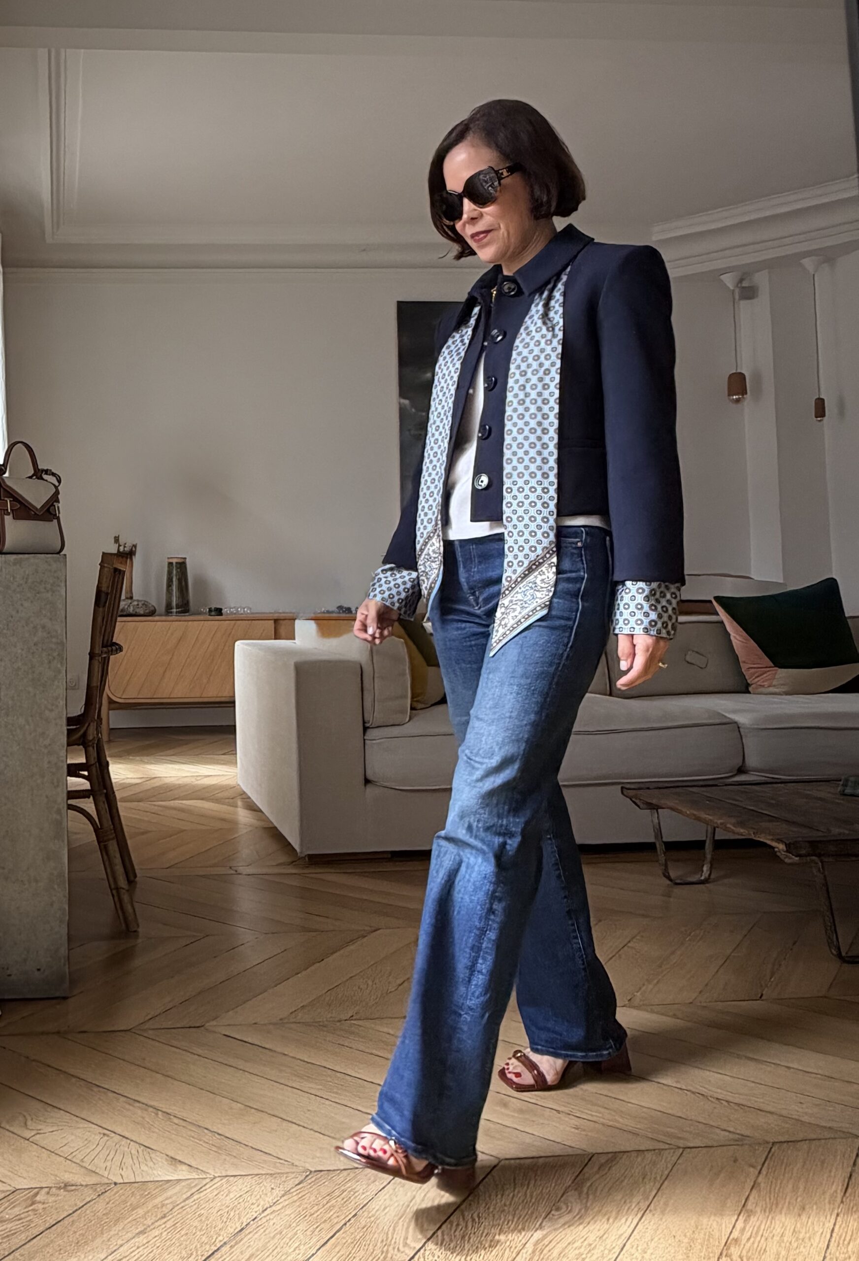 Woman in navy blazer with patterned silk scarf styled over flare jeans and strappy sandals in Paris apartment with herringbone floors.