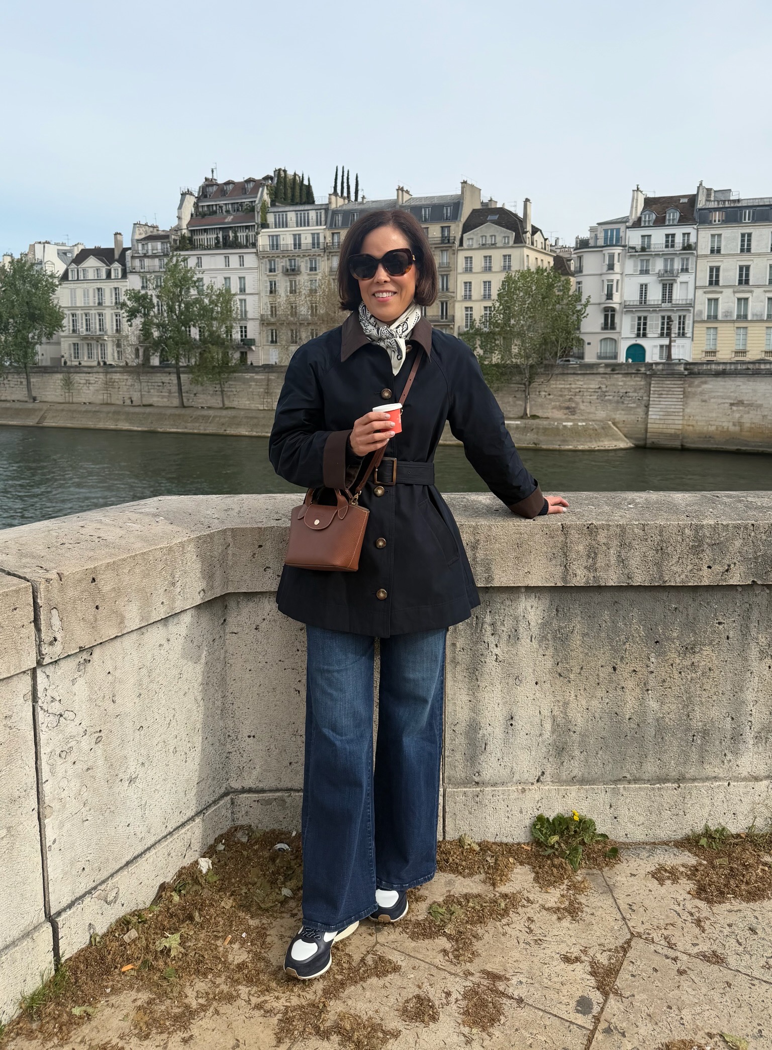 Woman in navy belted jacket, wide-leg jeans, and Longchamp mini bag standing along the Seine River in Paris/