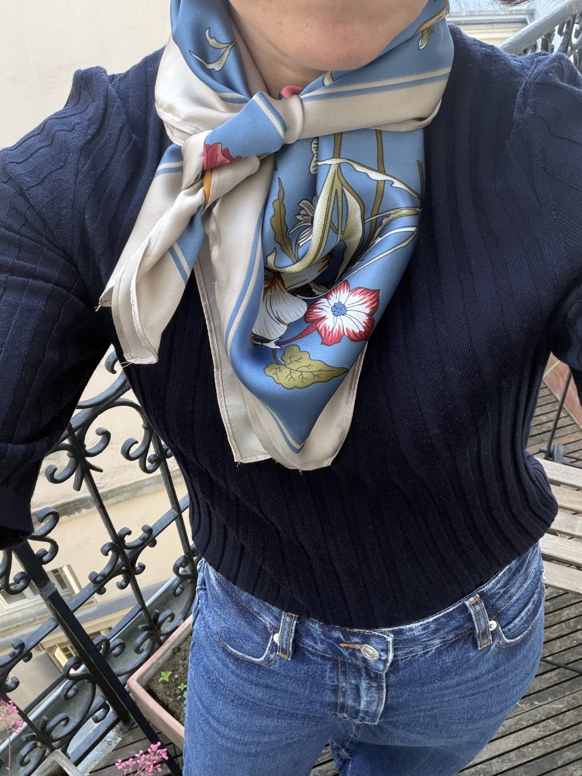 Close-up of a floral blue silk scarf tied at the neck over a navy ribbed sweater and jeans on a Paris balcony.