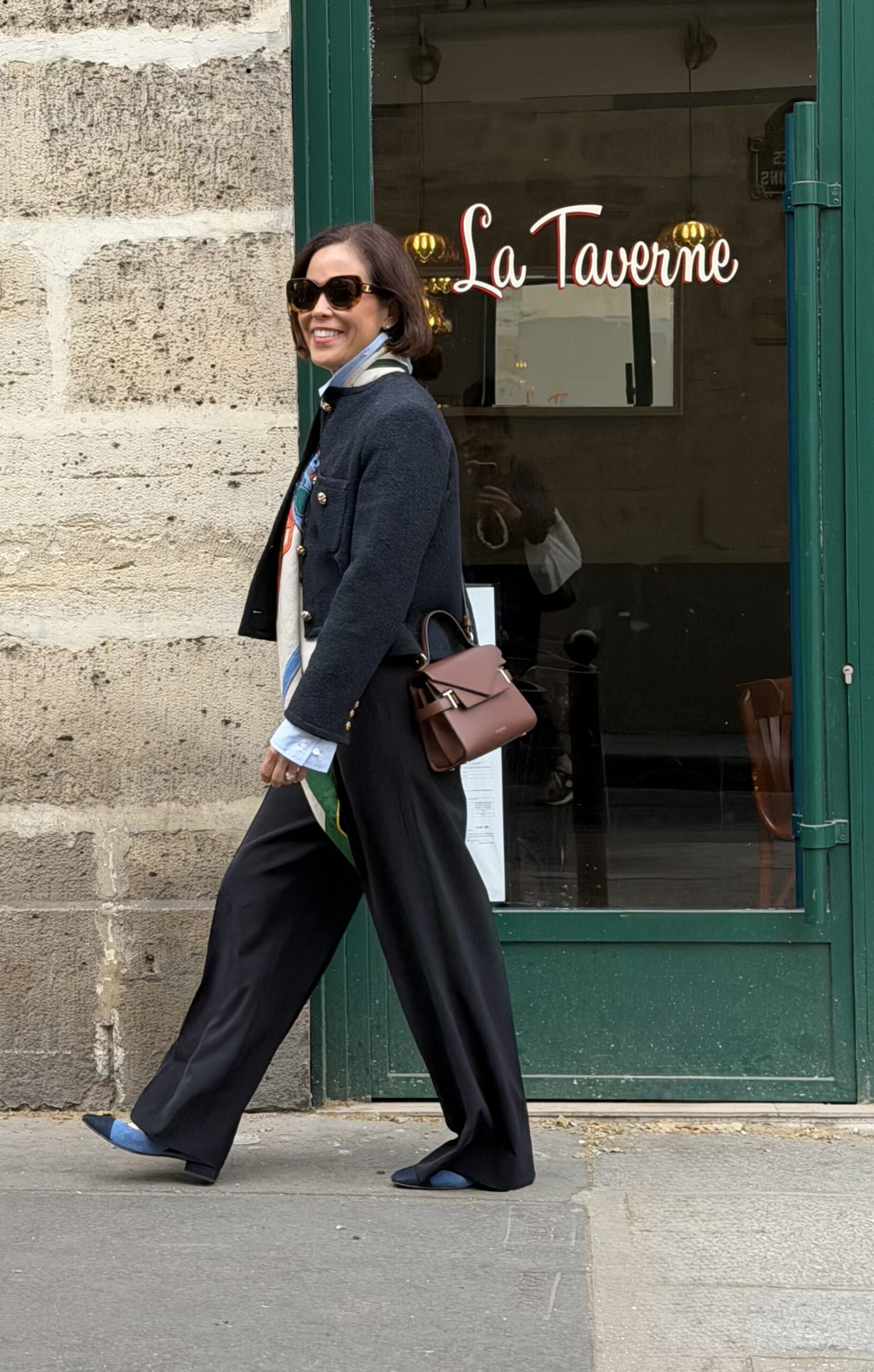 Woman in navy military-style jacket, black wide-leg trousers, and silk scarf walking past La Taverne bistro in Paris.