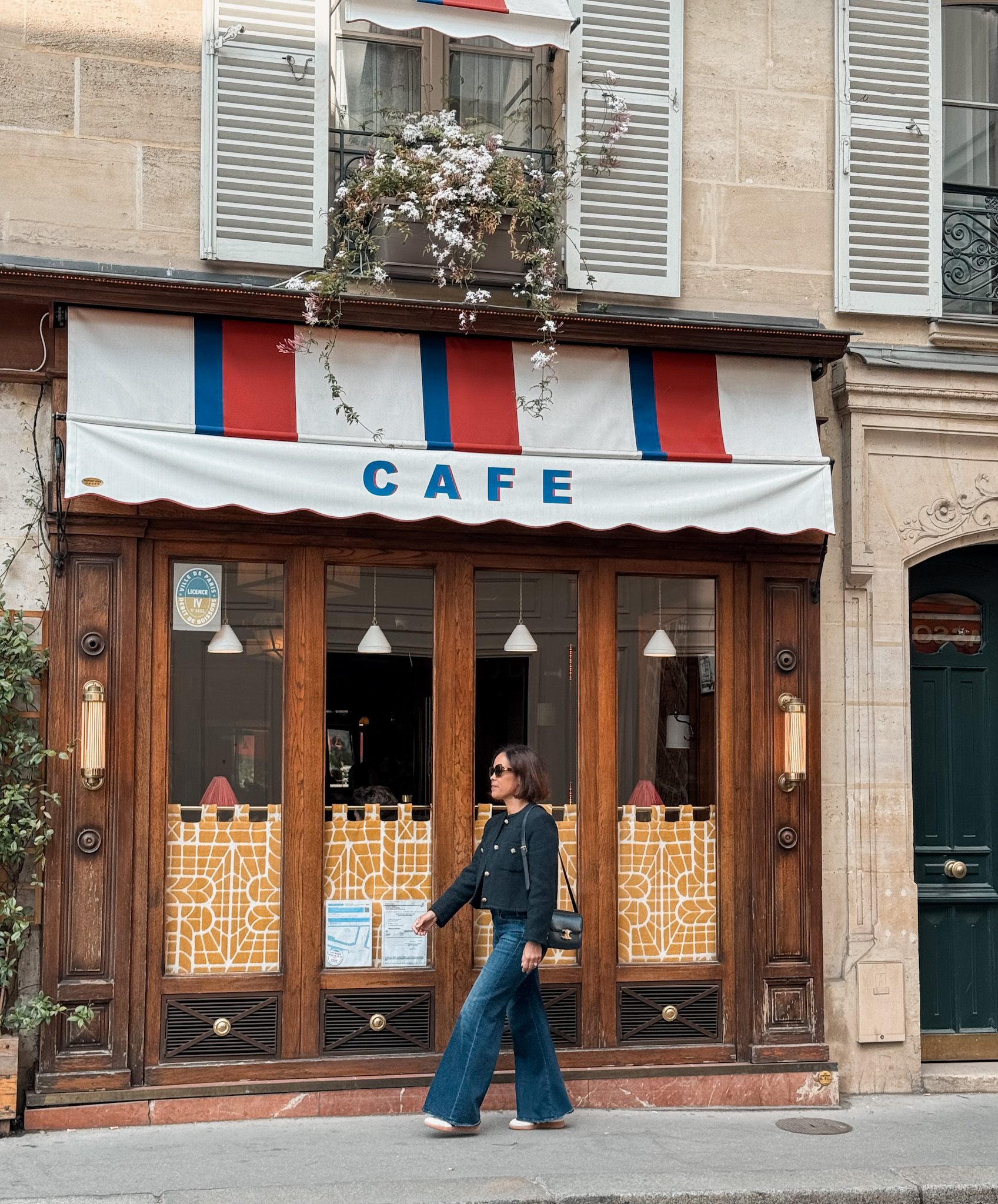 Woman walking past a classic Parisian café with red, white, and blue awning wearing a navy jacket, wide-leg jeans, and sneakers — effortless Paris street style