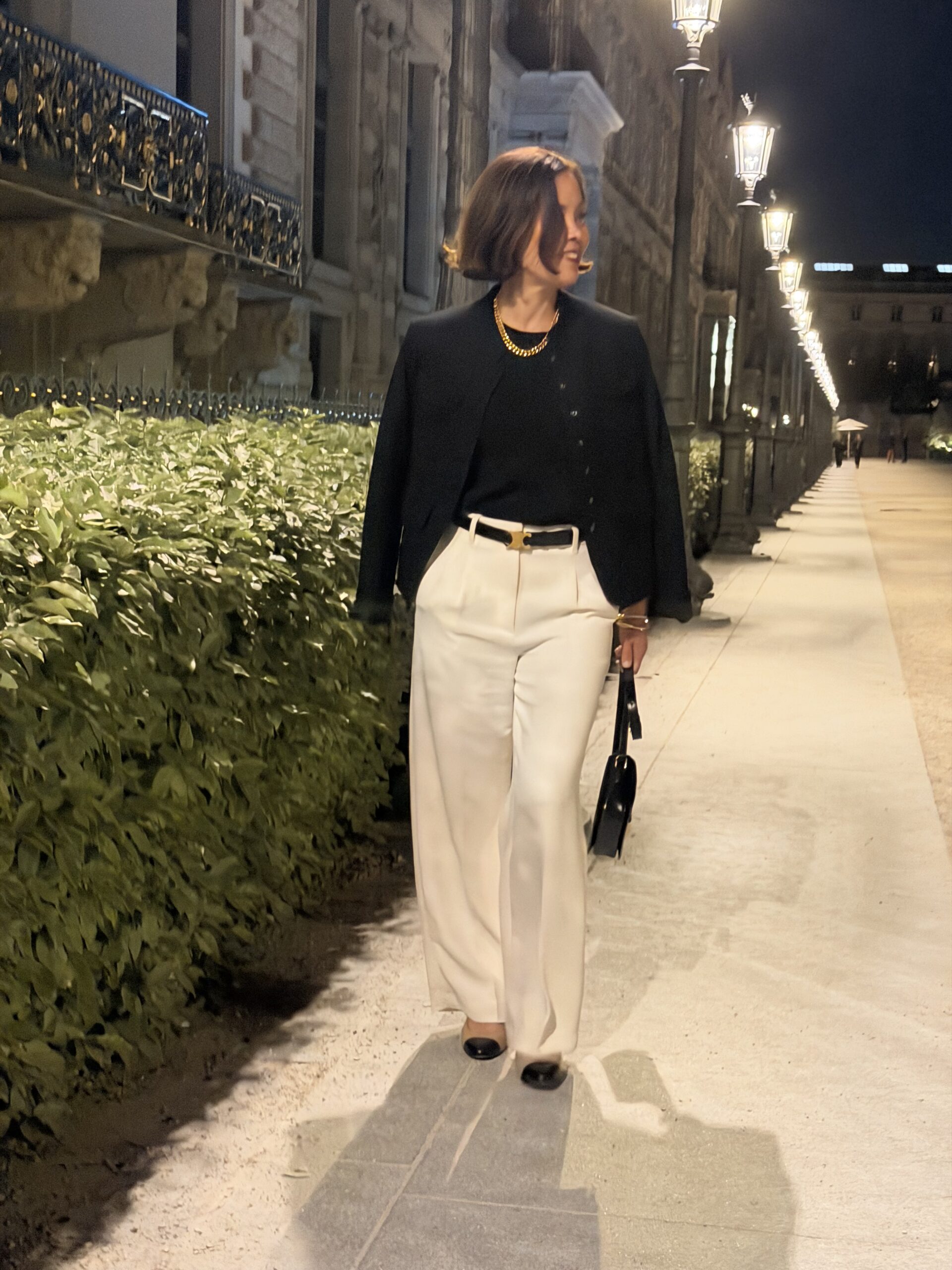 Woman in black blazer, ivory wide-leg trousers, white belt, and gold chain necklace walking Paris streets at night.