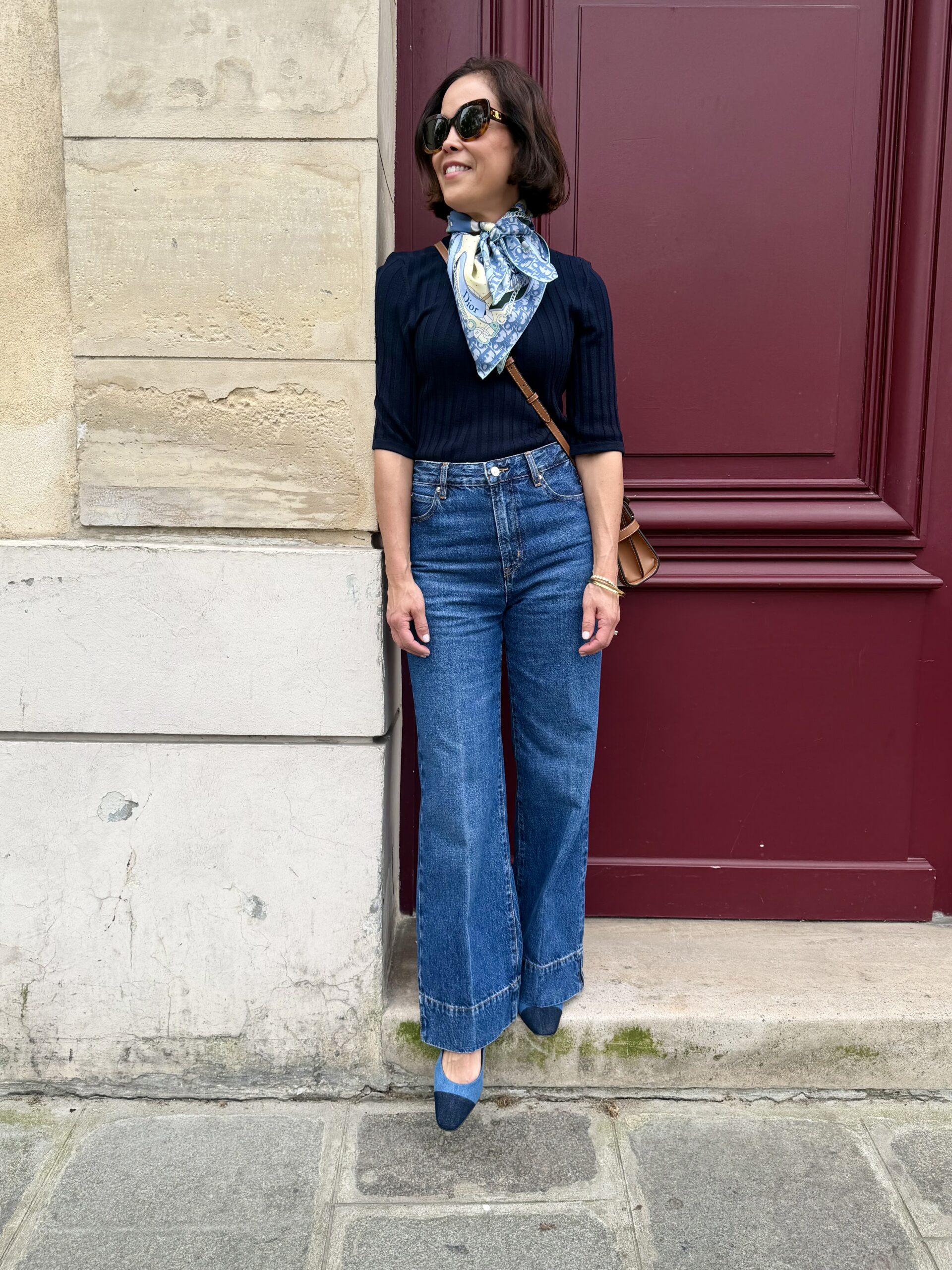 Woman in navy ribbed puff-sleeve sweater, wide-leg jeans, and Dior silk scarf leaning against a burgundy door in Paris.