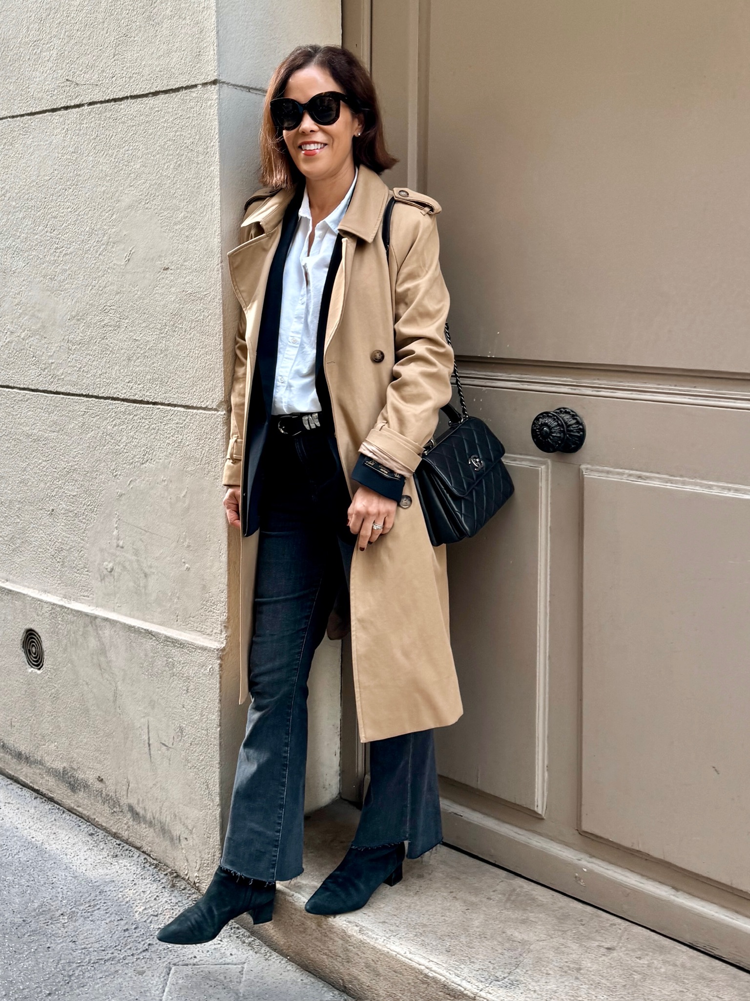 Woman in Paris wearing a camel trench coat, white button-down, dark jeans, black ankle boots, and quilted black handbag — a classic neutral Paris outfit