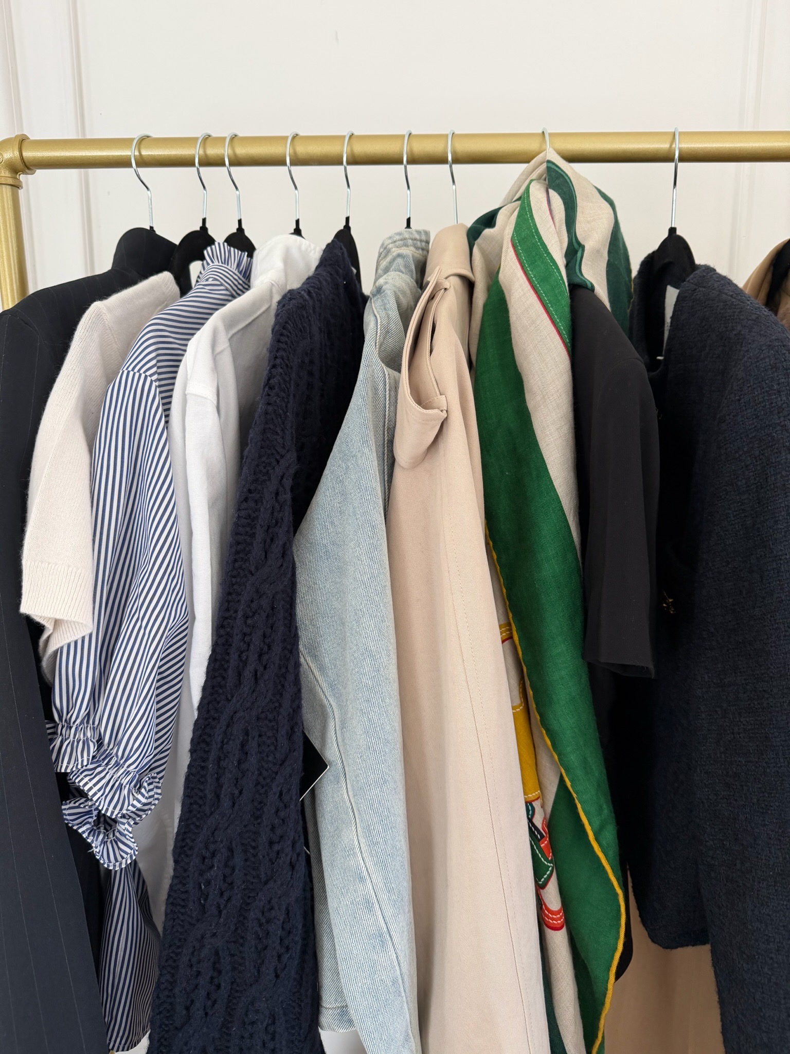 Neutral and timeless Paris travel outfits hanging on a gold clothing rack — navy blazer, striped shirt, white button-down, denim, and camel pieces pre-planned before packing