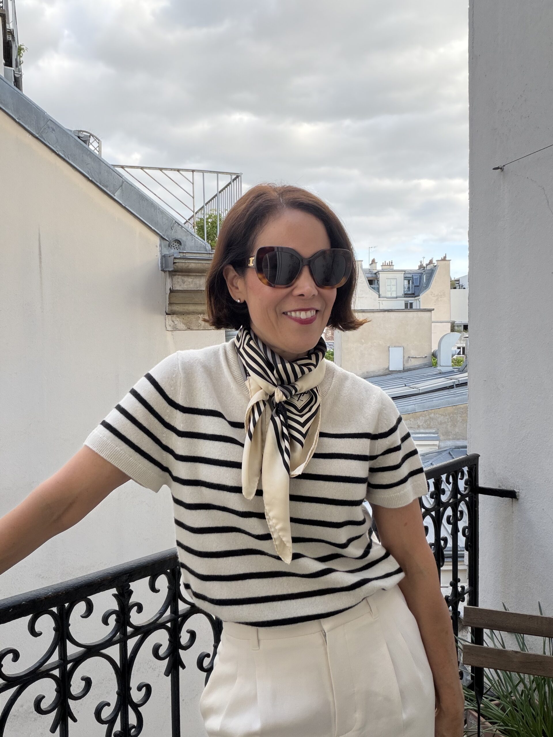 Woman in cream and black striped short-sleeve sweater with striped silk scarf tied at neck standing on Paris rooftop.