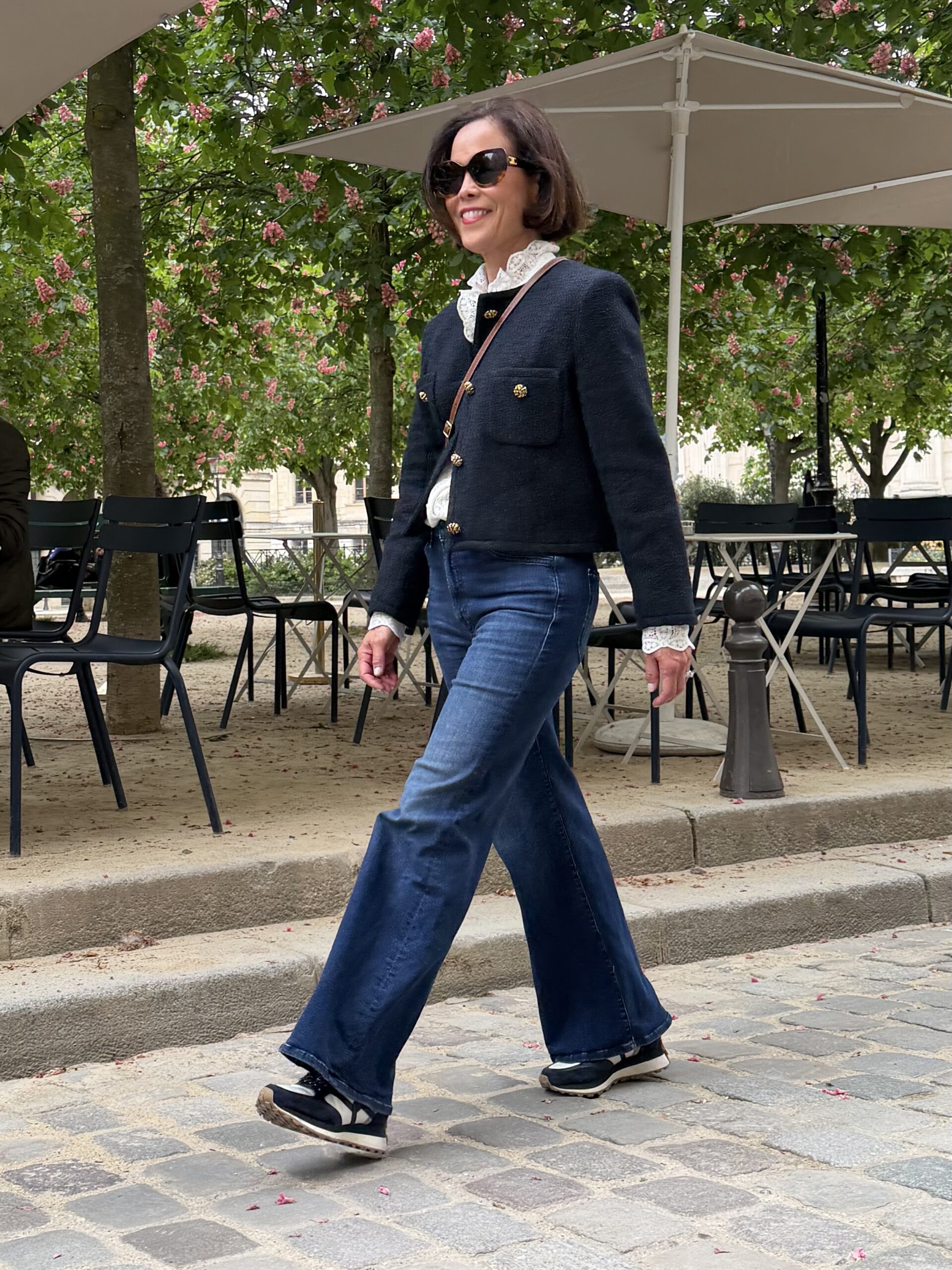 Woman wearing navy tweed jacket with gold buttons, lace blouse, and wide-leg jeans walking through Place Dauphne in Paris