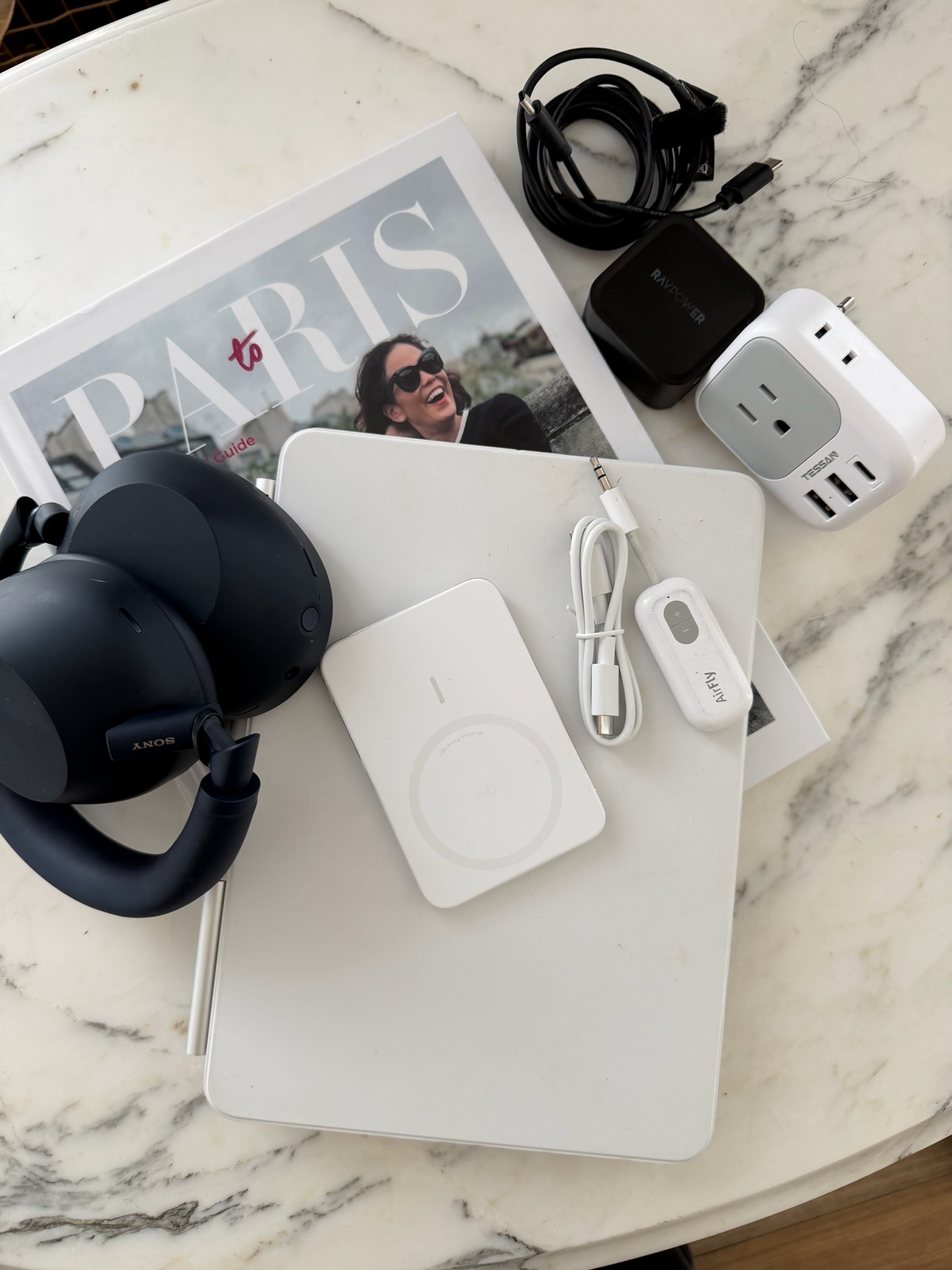 Paris travel tech essentials laid out on marble including Sony headphones, AirFly adapter, portable battery, EU power adapter, charging cables, and Essential Guide to Paris