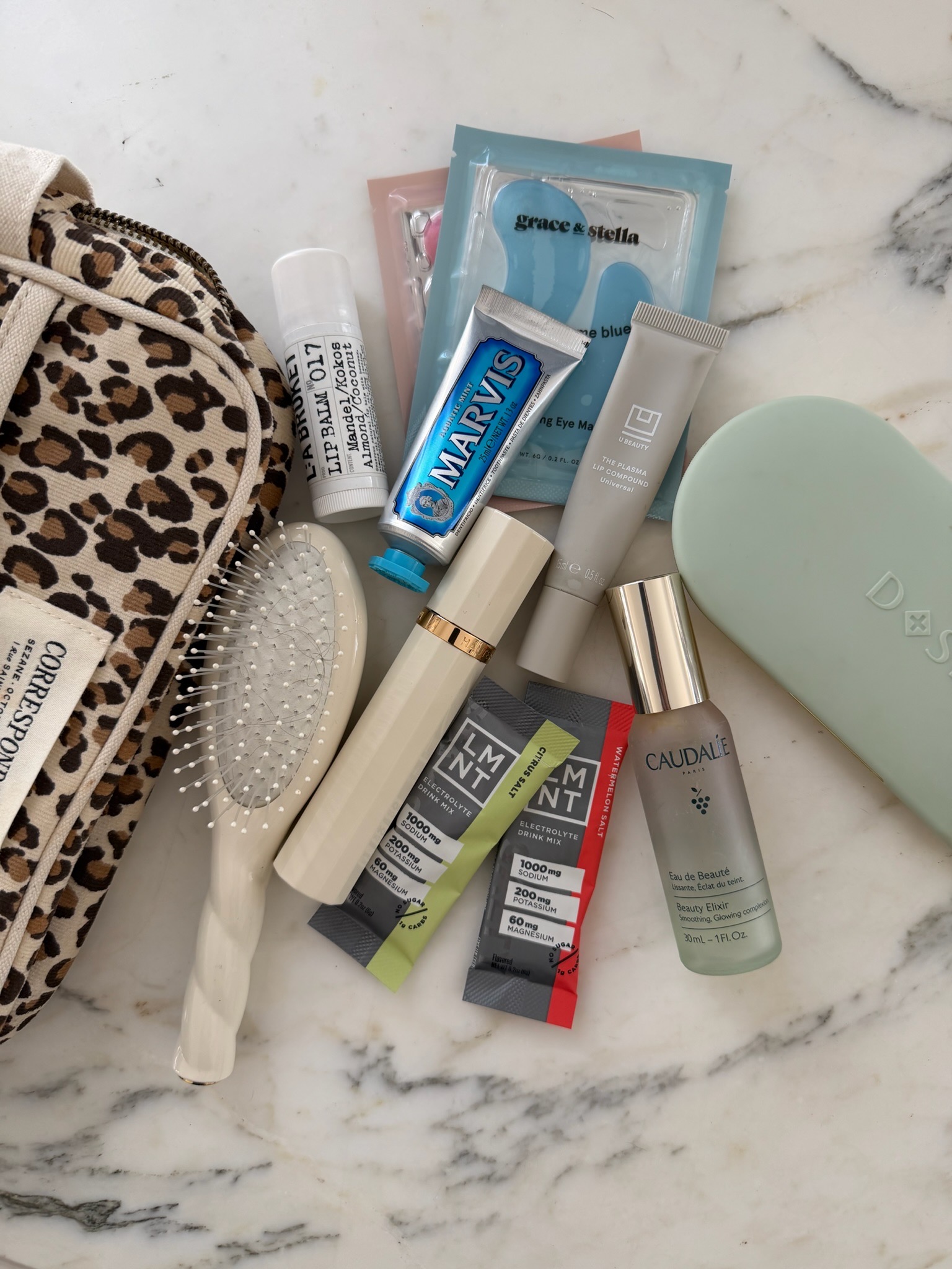 Flat lay of travel wellness pouch essentials including Marvis toothpaste, Caudalie face mist, LMNT electrolytes, Grace and Stella eye patches, lip balm, and mini hairbrush on marble
