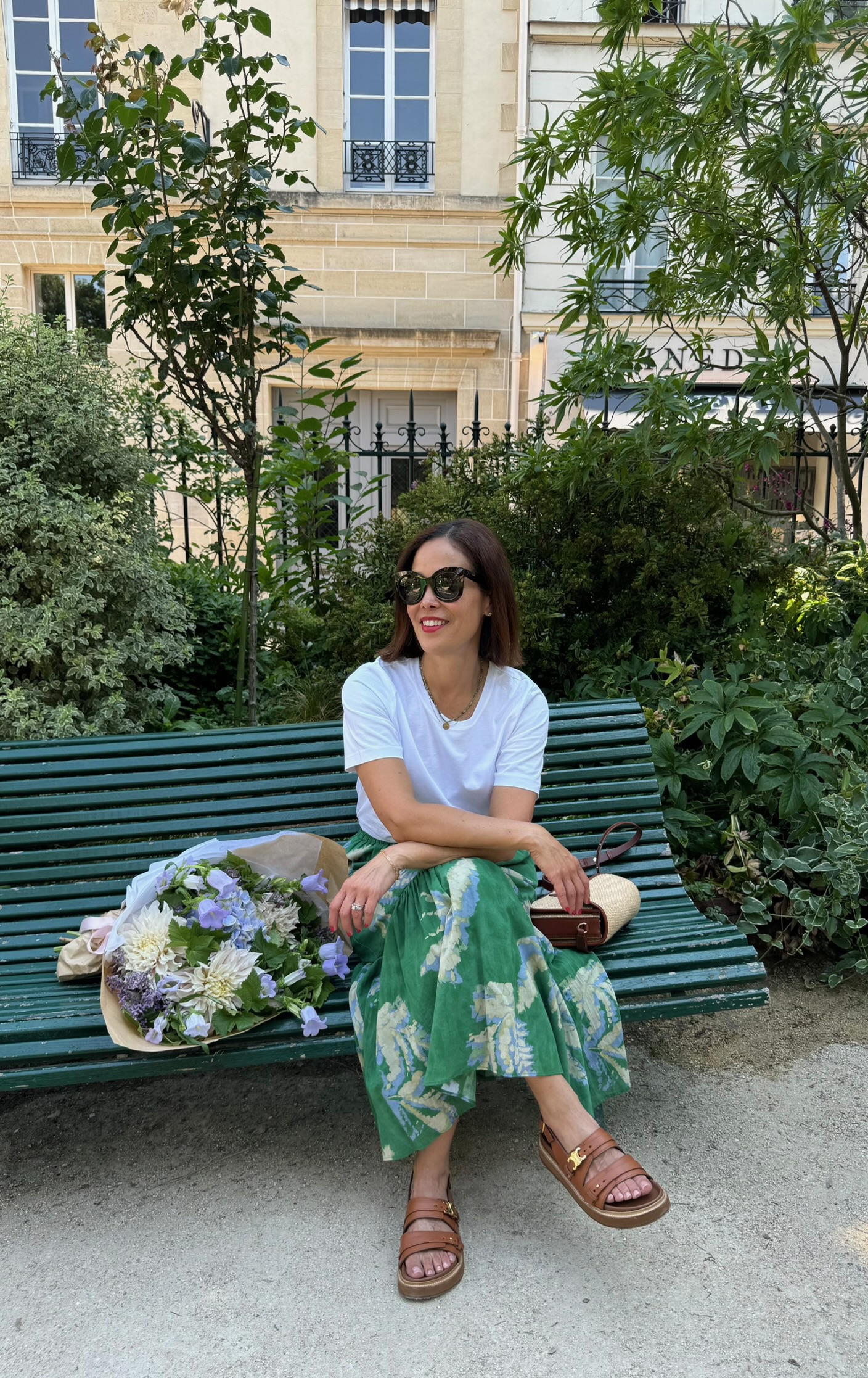 Woman in a white classic tee and green floral midi skirt with tan sandals and a basket bag sitting on a bench in a Paris garden with a bouquet of flowers