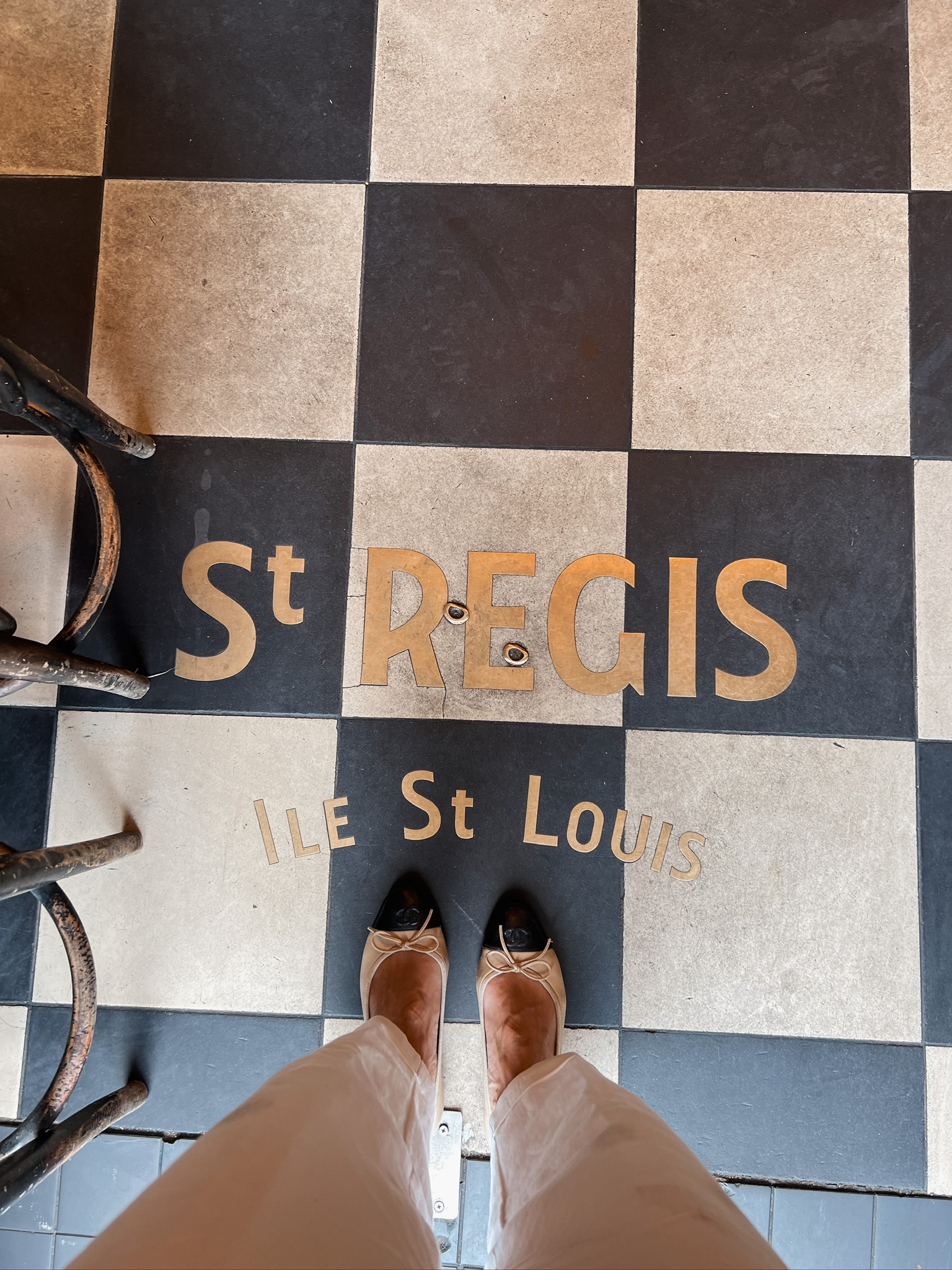 Tan ballet flats with bow detail worn with beige wide-leg trousers on the checkered tile floor of Cafe St Regis on Ile Saint Louis in Paris