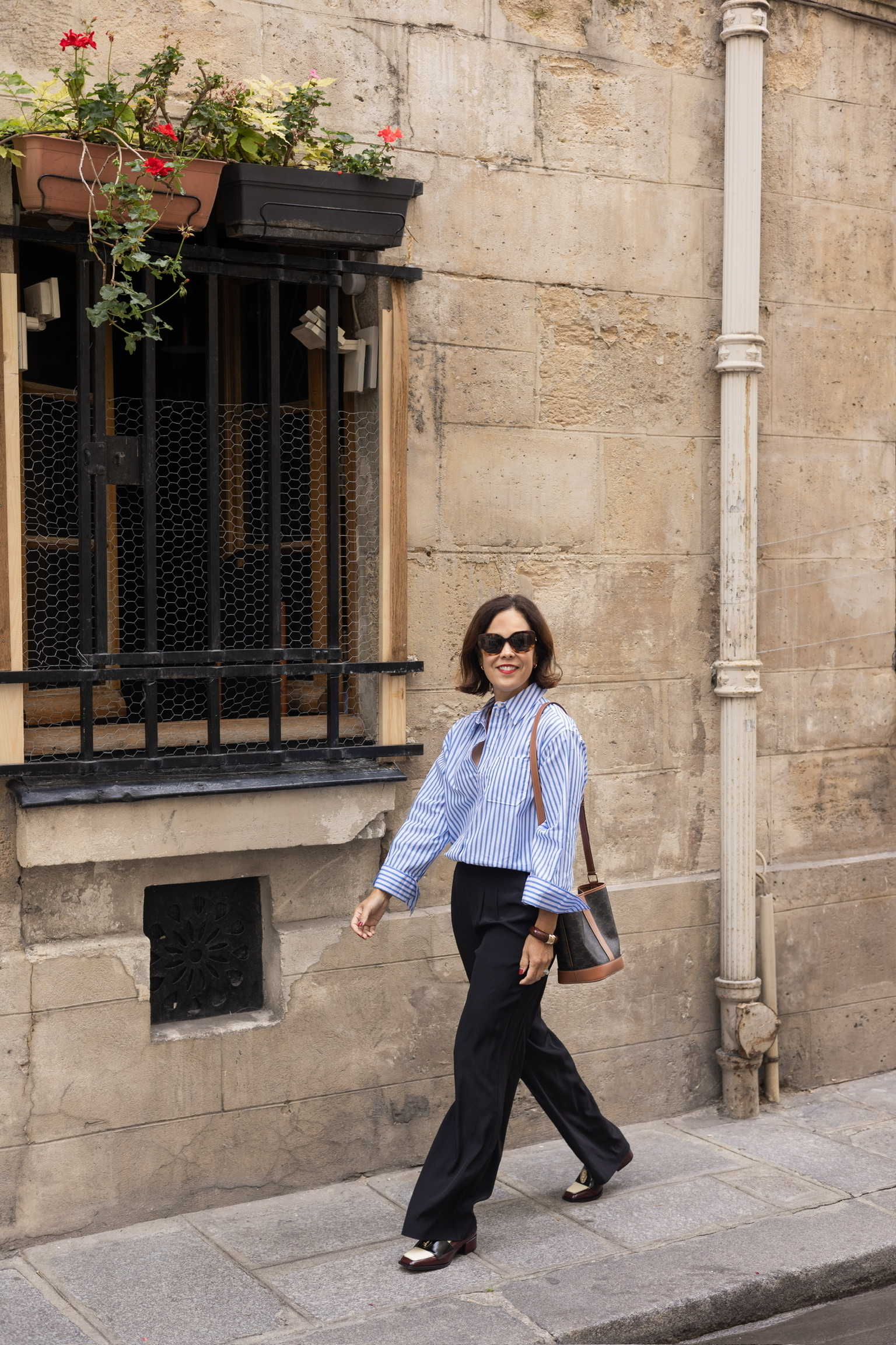 Woman in a blue and white striped cotton blouse with wide-leg black trousers and two-tone loafers walking a cobblestone street in Paris