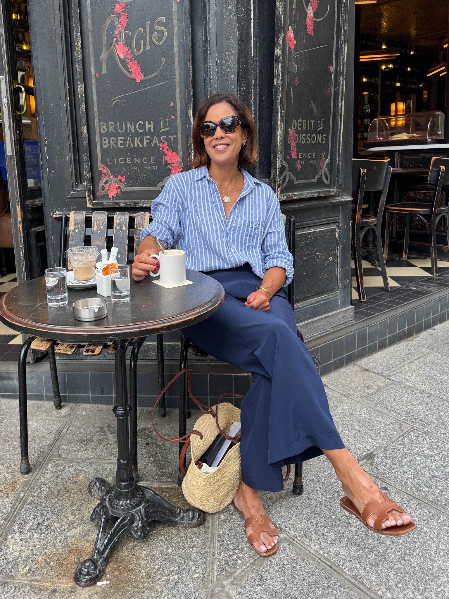 Woman in a blue striped linen shirt and navy wide-leg trousers with tan slide sandals and a basket bag sitting at an outdoor cafe table in Paris