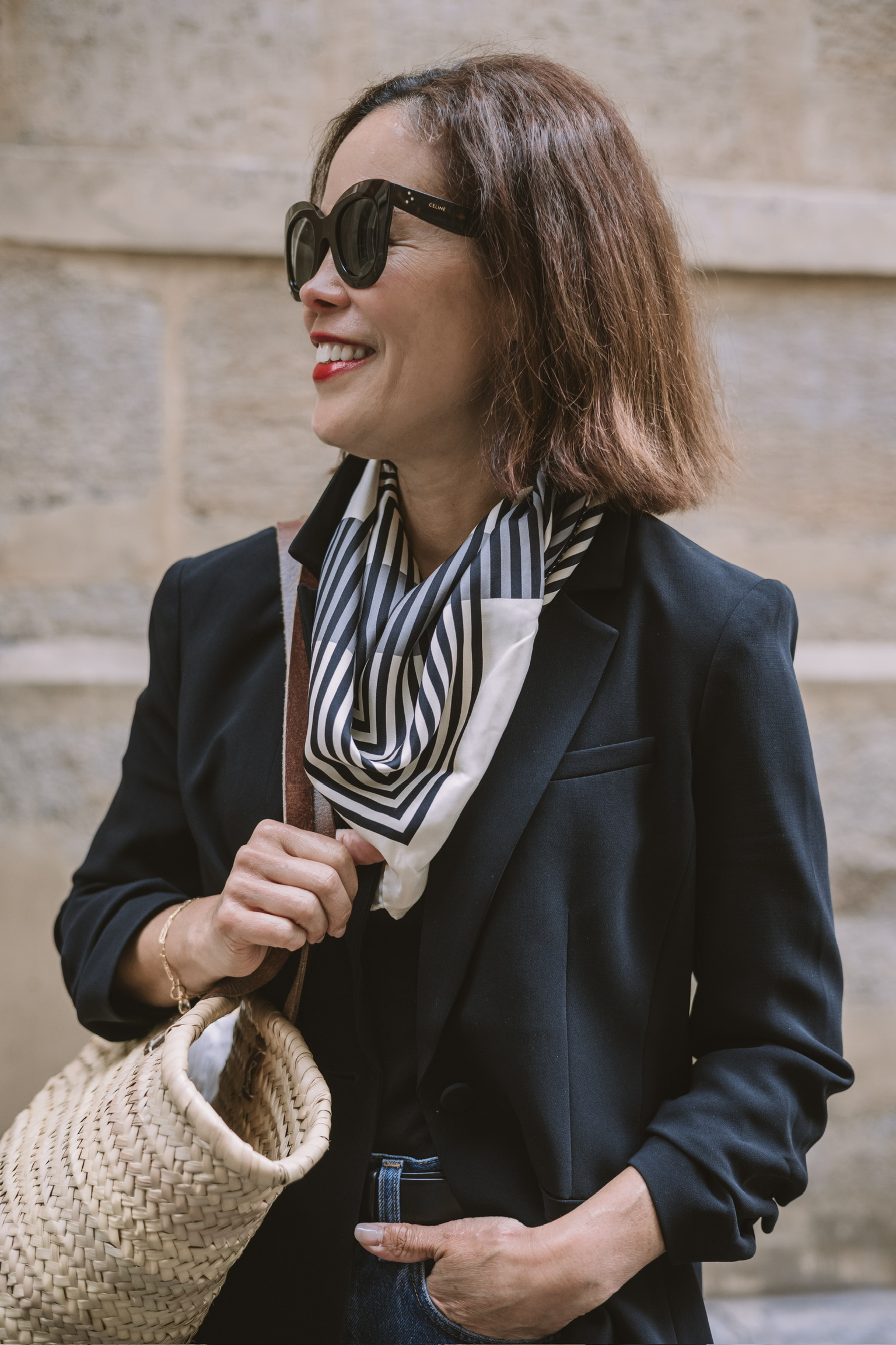 Woman over 40 in black blazer with striped silk scarf and woven straw bag on Paris street — the blazer French capsule wardrobe