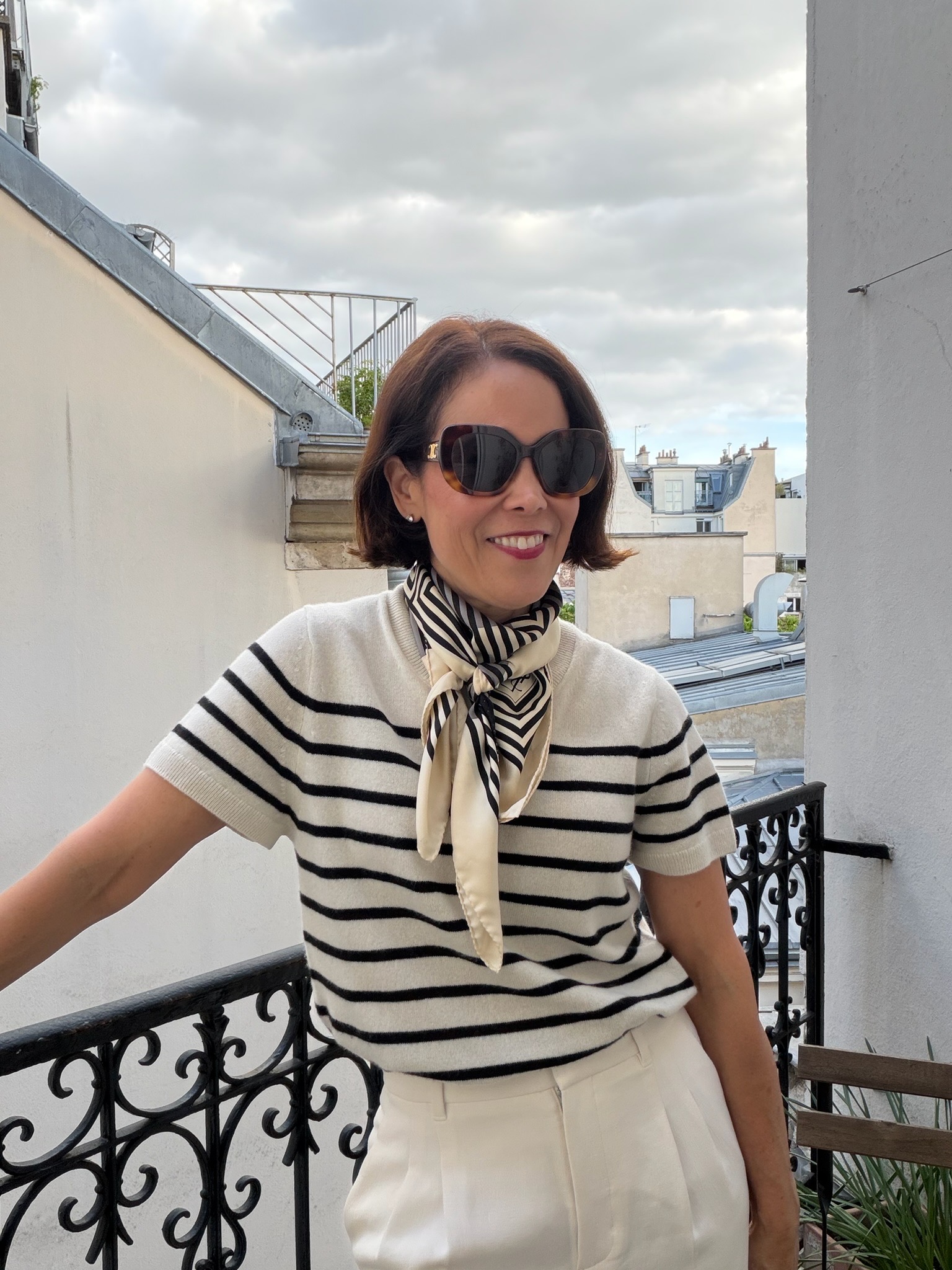 Woman in cream and black Breton stripe knit top with silk scarf tied at neck on Paris rooftop balcony — French capsule wardrobe women 40+