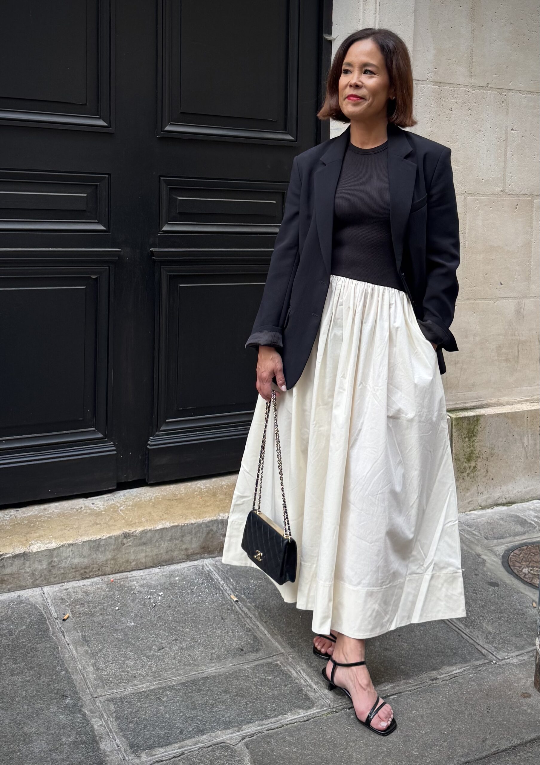 Woman in cream midi skirt with black blazer and ribbed knit top holding Chanel bag in front of black Parisian door — the dress French capsule wardrobe women 40+