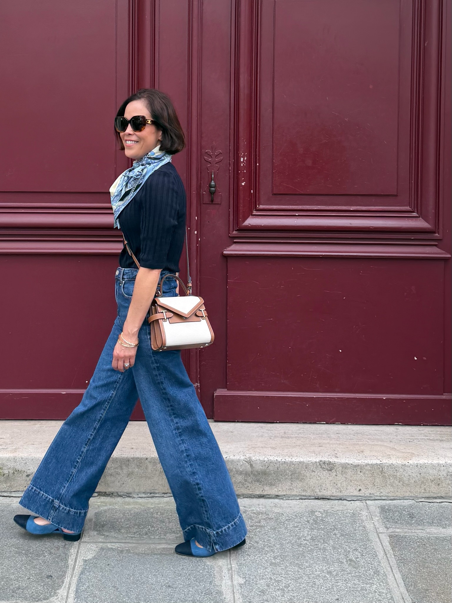 Woman in navy ribbed knit top tucked into wide-leg denim with silk scarf and structured mini bag against burgundy Paris door — lightweight knitwear French capsule wardrobe