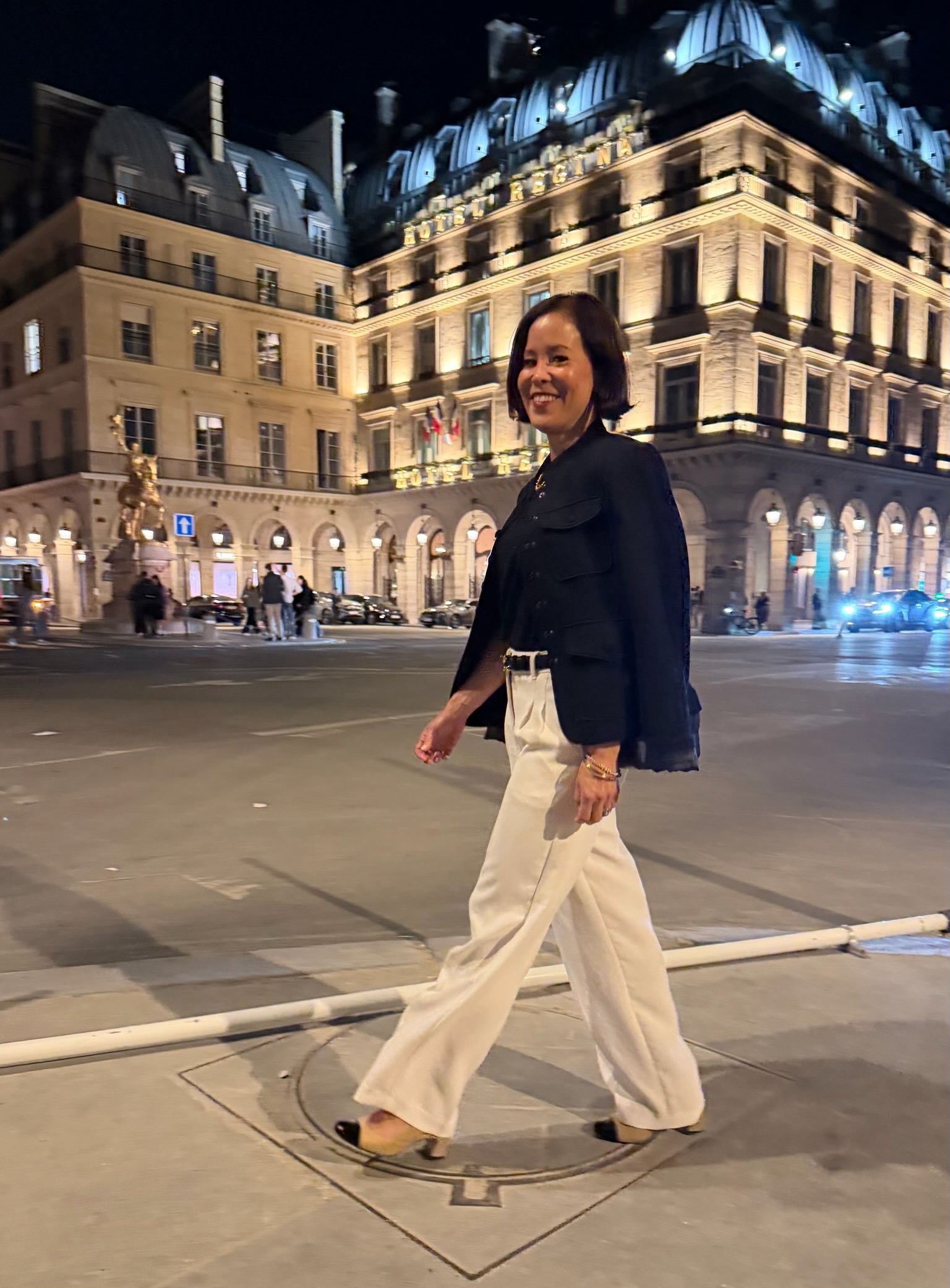 Woman in white wide-leg tailored trousers and navy blazer walking at night in Paris near Hotel Regina — tailored trouser French capsule wardrobe