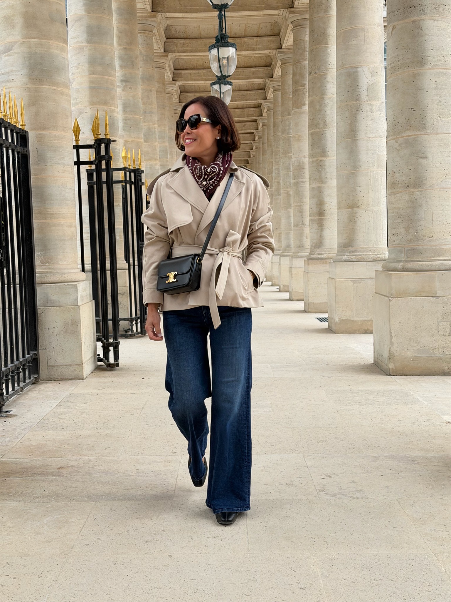 Woman in wide-leg jeans, trench coat, and bandana scarf walking through Palais Royal arcades Paris — French capsule wardrobe