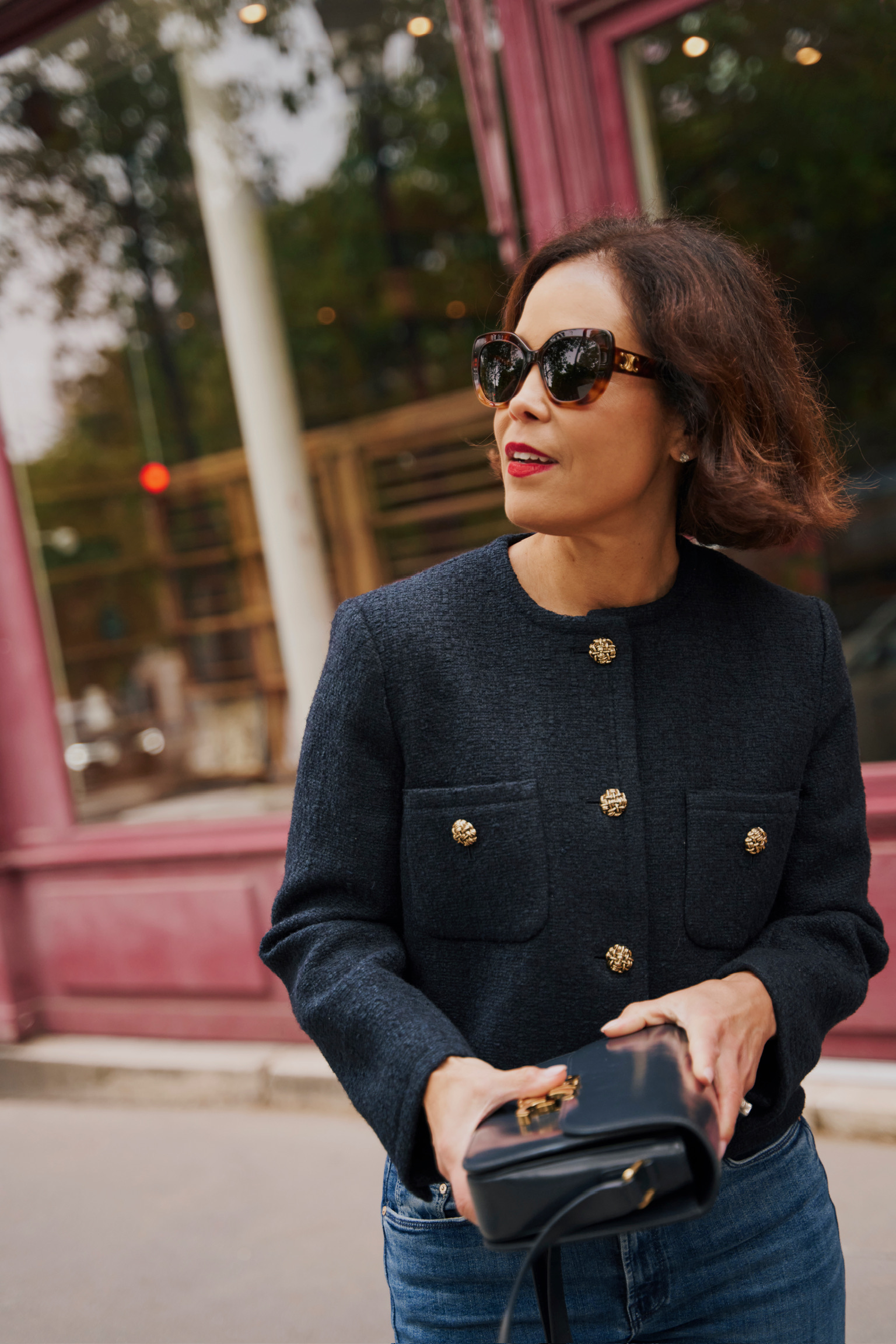 Woman over 40 wearing navy tweed jacket with gold buttons and structured black bag on Paris street — French capsule wardrobe