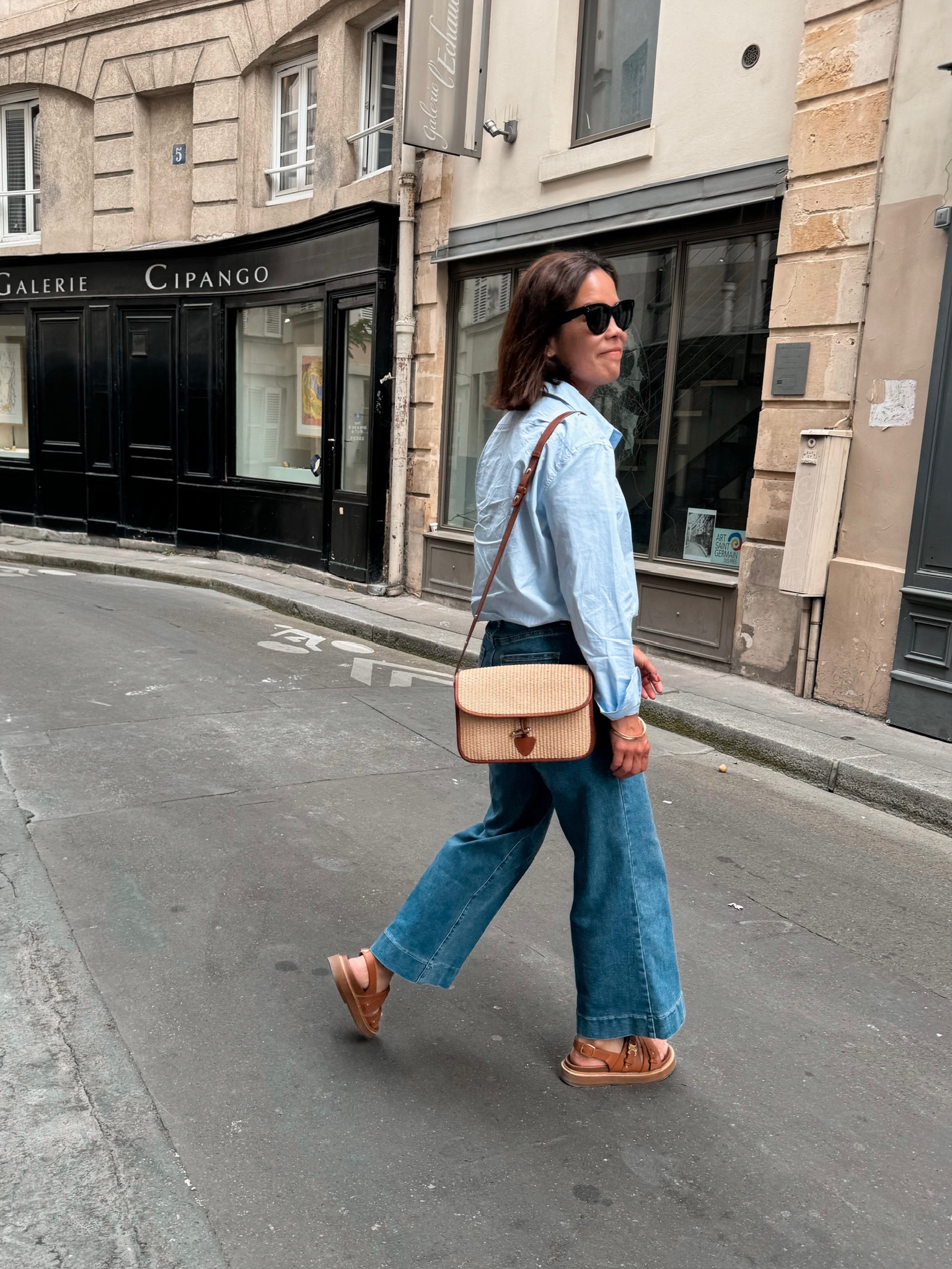 Woman in light wash wide-leg denim jeans and a pale blue linen shirt with tan sandals and a woven crossbody bag walking a Paris street