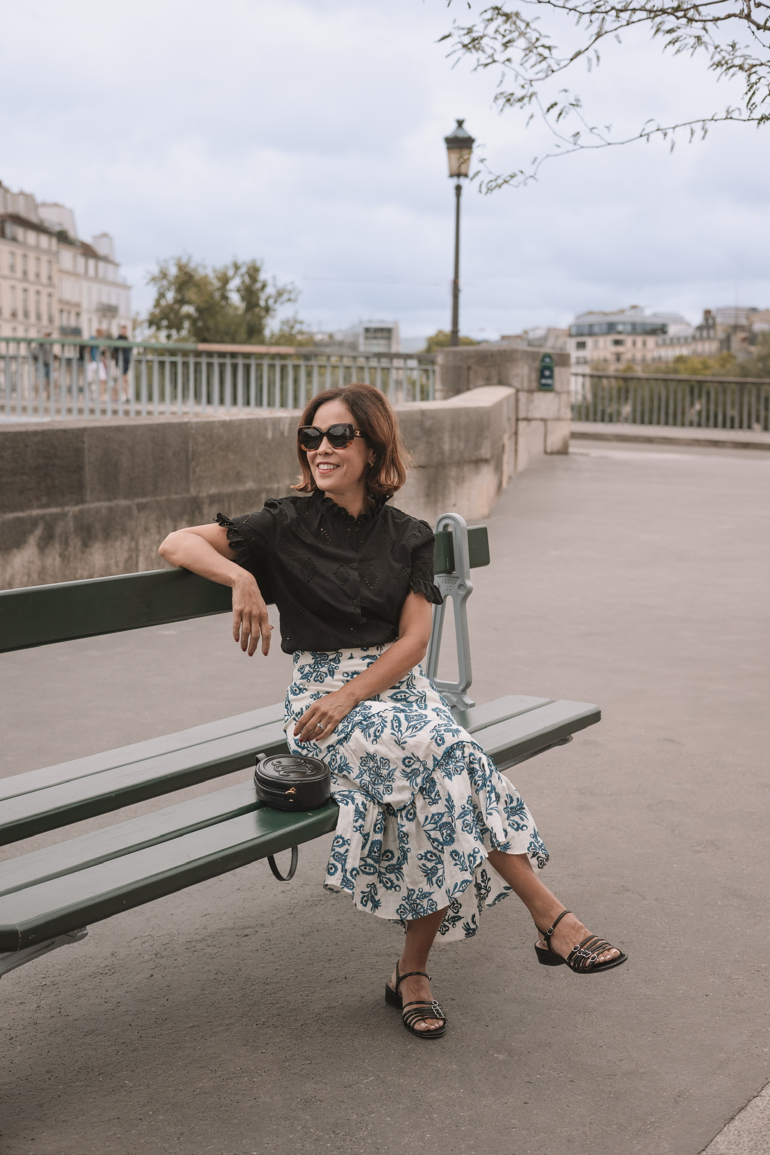 Woman in black strappy sandals with a blue and white floral midi skirt and black eyelet top sitting on a green Paris bench along the Seine