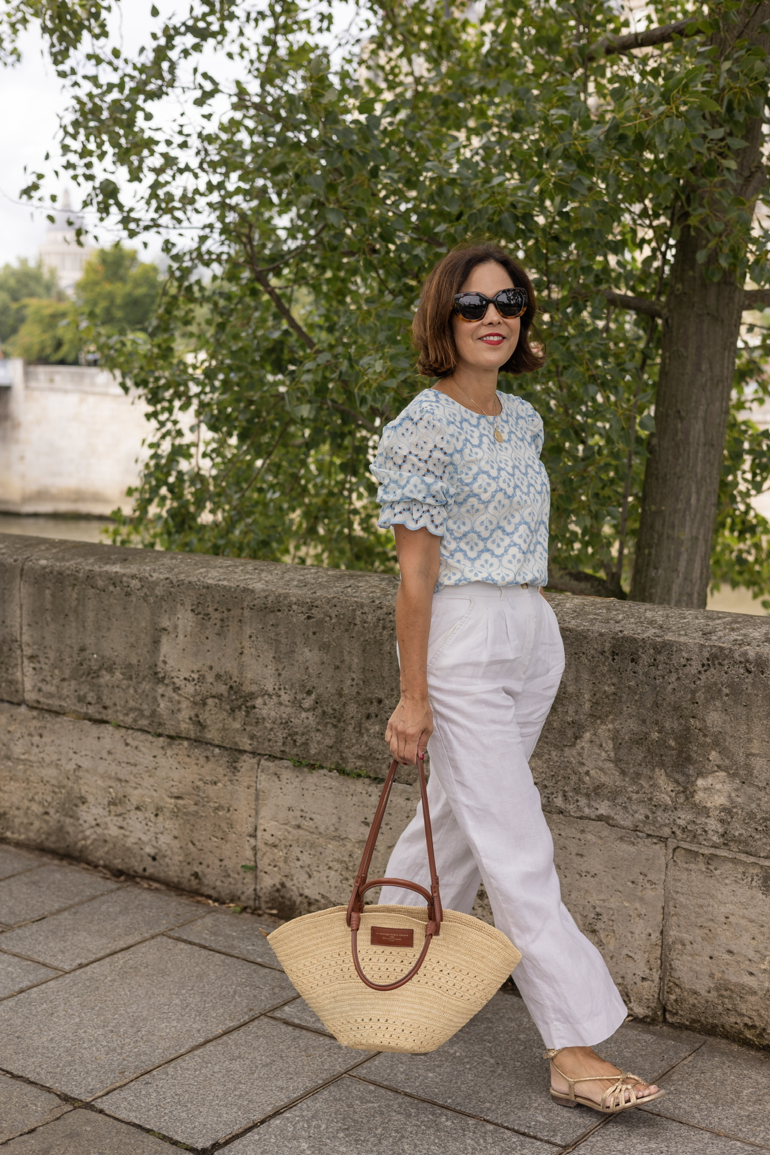 Woman in white wide-leg linen trousers and a blue eyelet top with a woven basket bag and strappy sandals walking along the Seine River in Paris