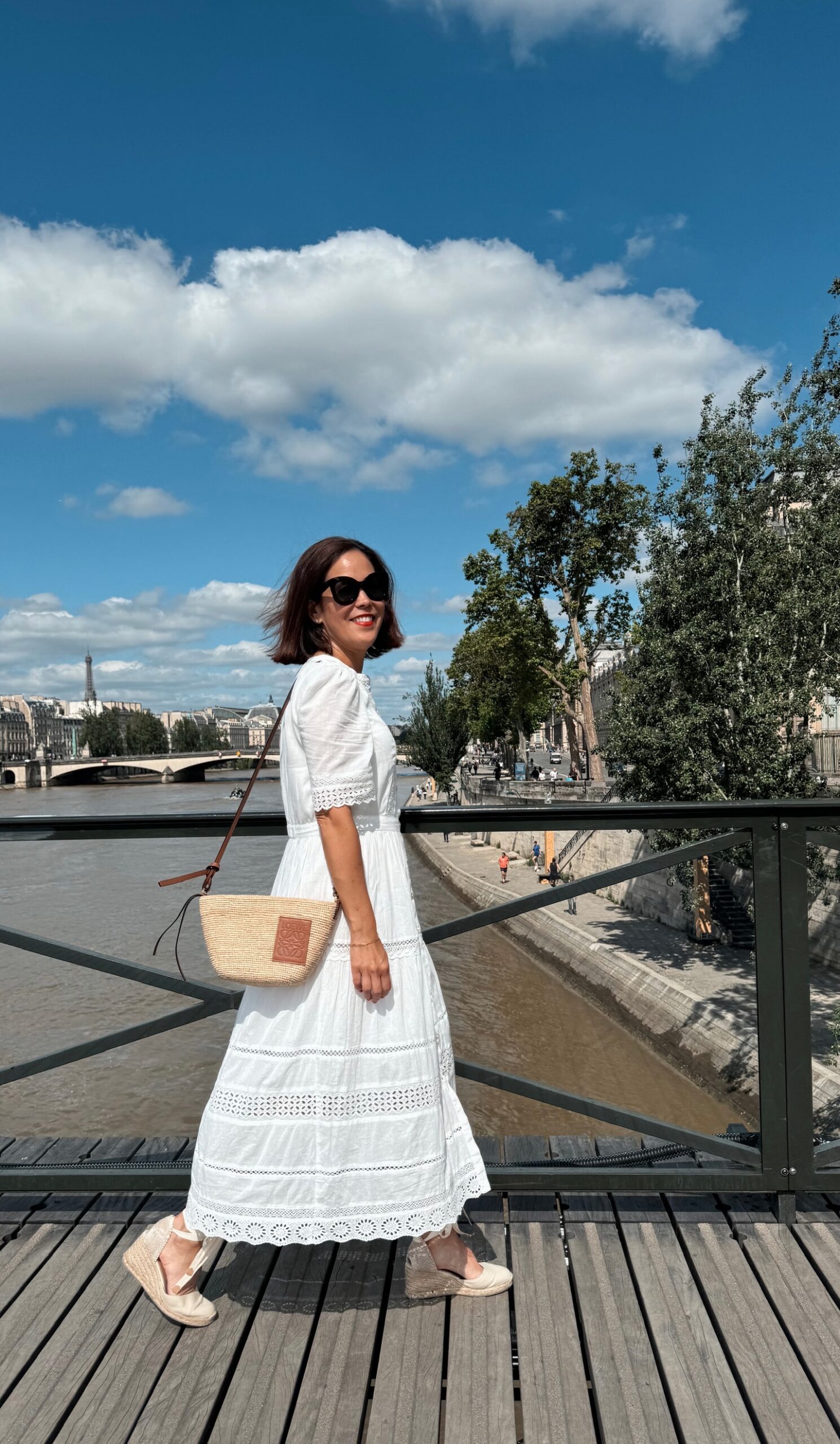 Woman in white eyelet midi dress with basket bag and espadrilles standing on a bridge over the Seine River in Paris with the Eiffel Tower in the background
